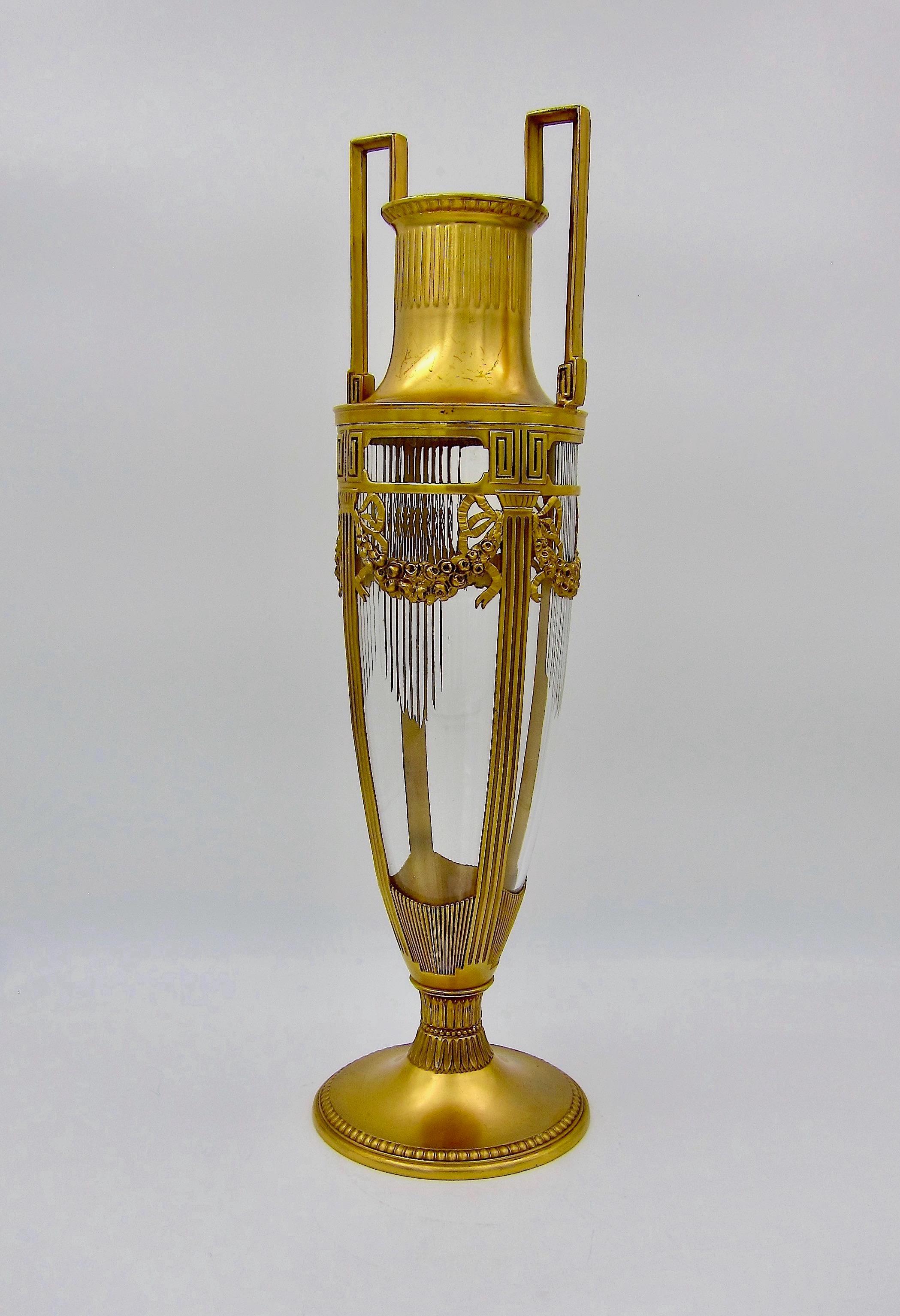A large antique vase in the neoclassical style with gilt metal mounts and a heavy colorless cut crystal glass insert. The tall and slender two-handled vase is an impressive 15.75 in. height and was created circa 1905 during the Art Nouveau /