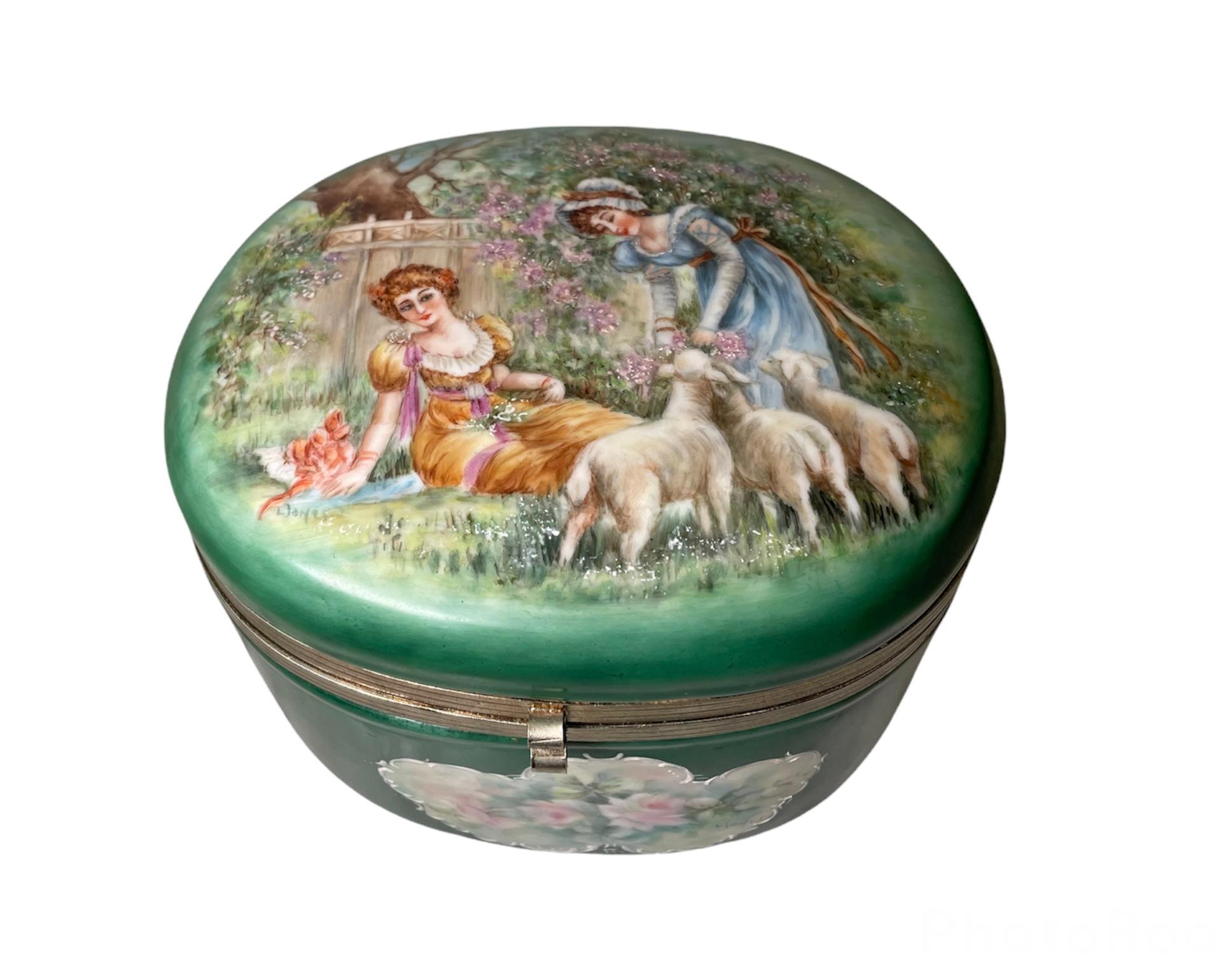 This a large oval hand painted porcelain vanity box. It depicts a scene of two Victorian young ladies in a garden. One of the ladies is seated in the green grass observing the other one that has some flowers in her hands while some lambs are trying