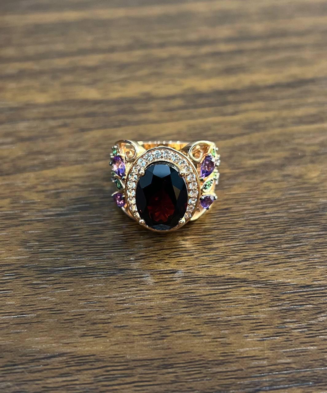 A striking multi-stone statement ring.
This exquisite piece features a large oval-cut dark red garnet at its center, surrounded by a halo of sparkling white diamonds. The intricate design includes vibrant purple amethyst and green tsavorites set