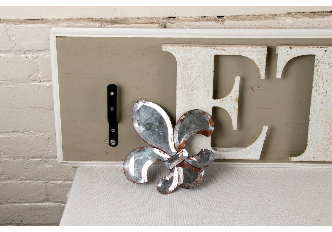 Un signe décoratif. En lettres capitales séparées en métal peint en blanc avec des motifs de fleurs de lys en miroir appliqués aux extrémités sur un fond de bois peint en gris. 
Dimensions : 74