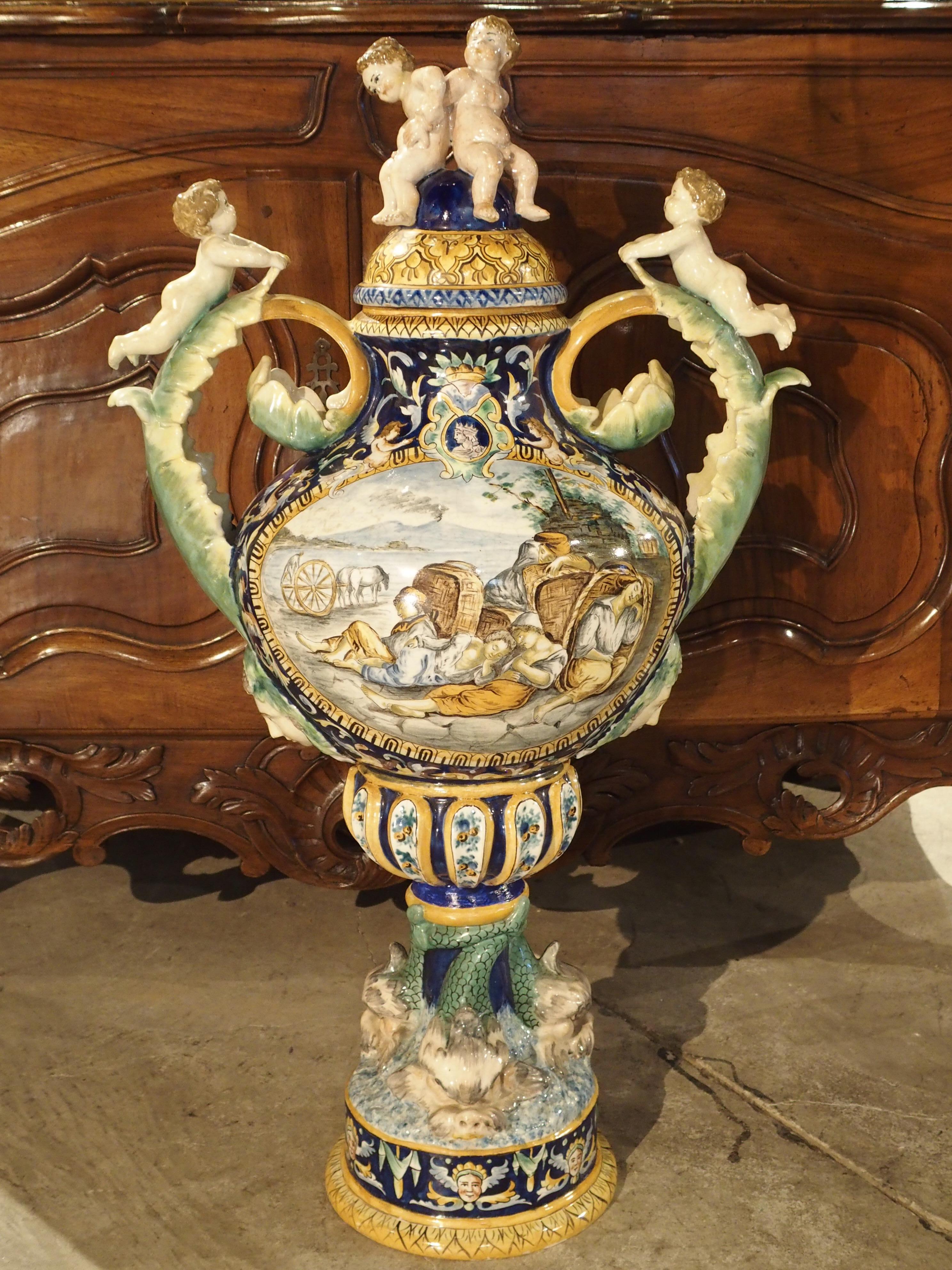From Italy, this large, hand-painted Majolica urn dates to the late 1800s. At the top are two putti seated upon the lid and two more putti lying upon their stomachs on the curled acanthus leaf handles. Two detailed scenes in cartouches have been