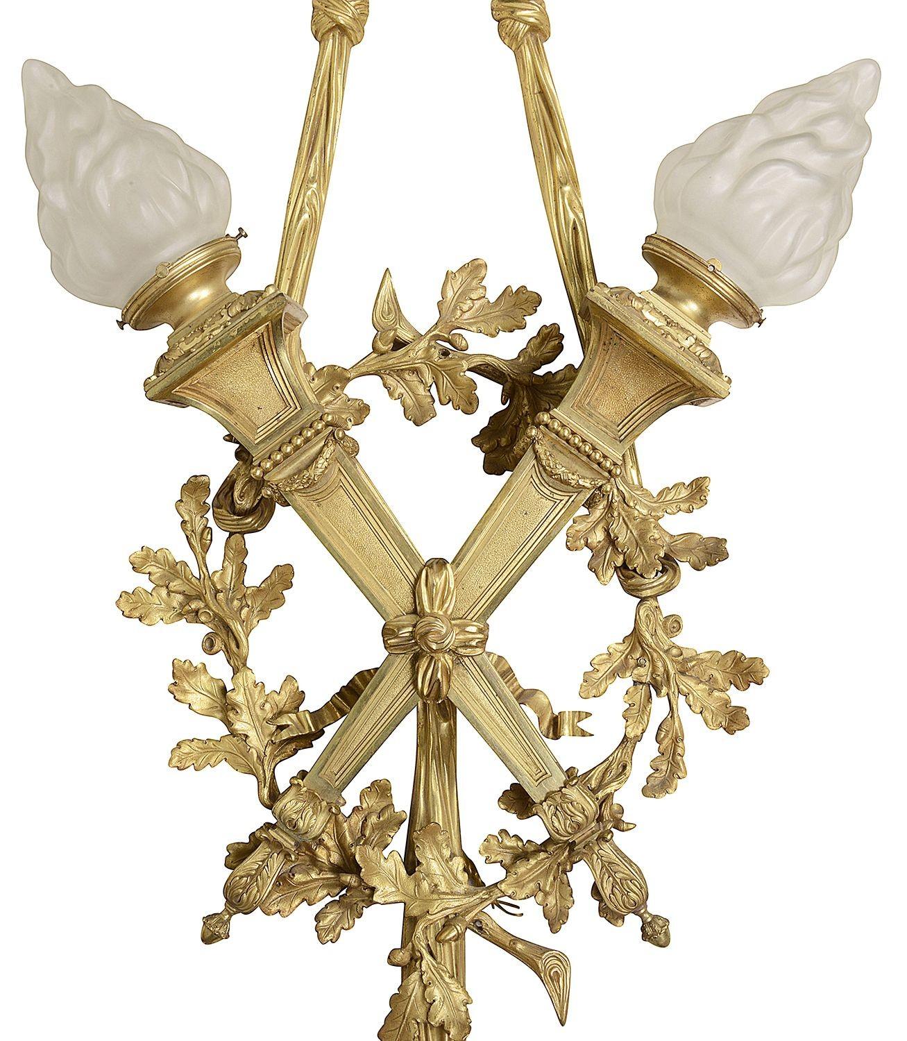 A large and impressive pair of classical Louis XVI style gilded ormolu wall lights, with flamed frosted glass shades on torches, held by a ribbon and bow with foliate wreaths to each.