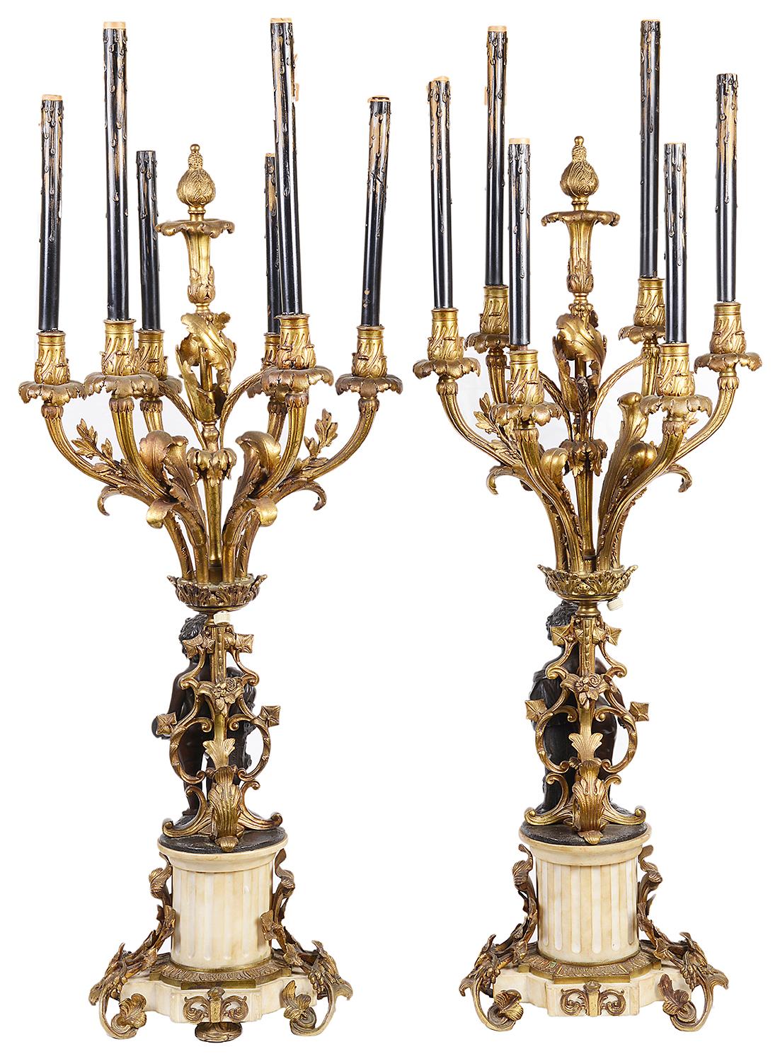 An impressive pair of 19th century French gilded ormolu, bronze and white marble candelabra, each having six scrolling foliate branches with candle sconces, above a pair of bronze semi clad putti, on a sculptor the other an artist, raised on
