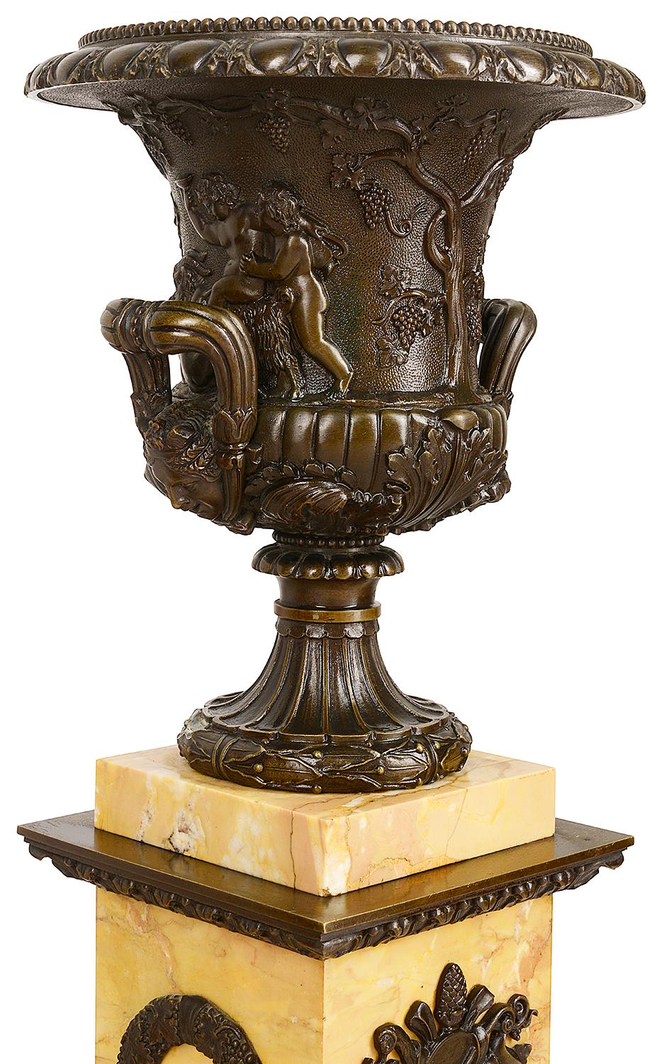 A striking pair of 19th century patinated bronze Capana shaped neoclassical bronze urns mounted on sienna marble plinths. Each with putti playing amongst grape vines, twin scrolling handles to each with egg and dart mould, foliate decoration and