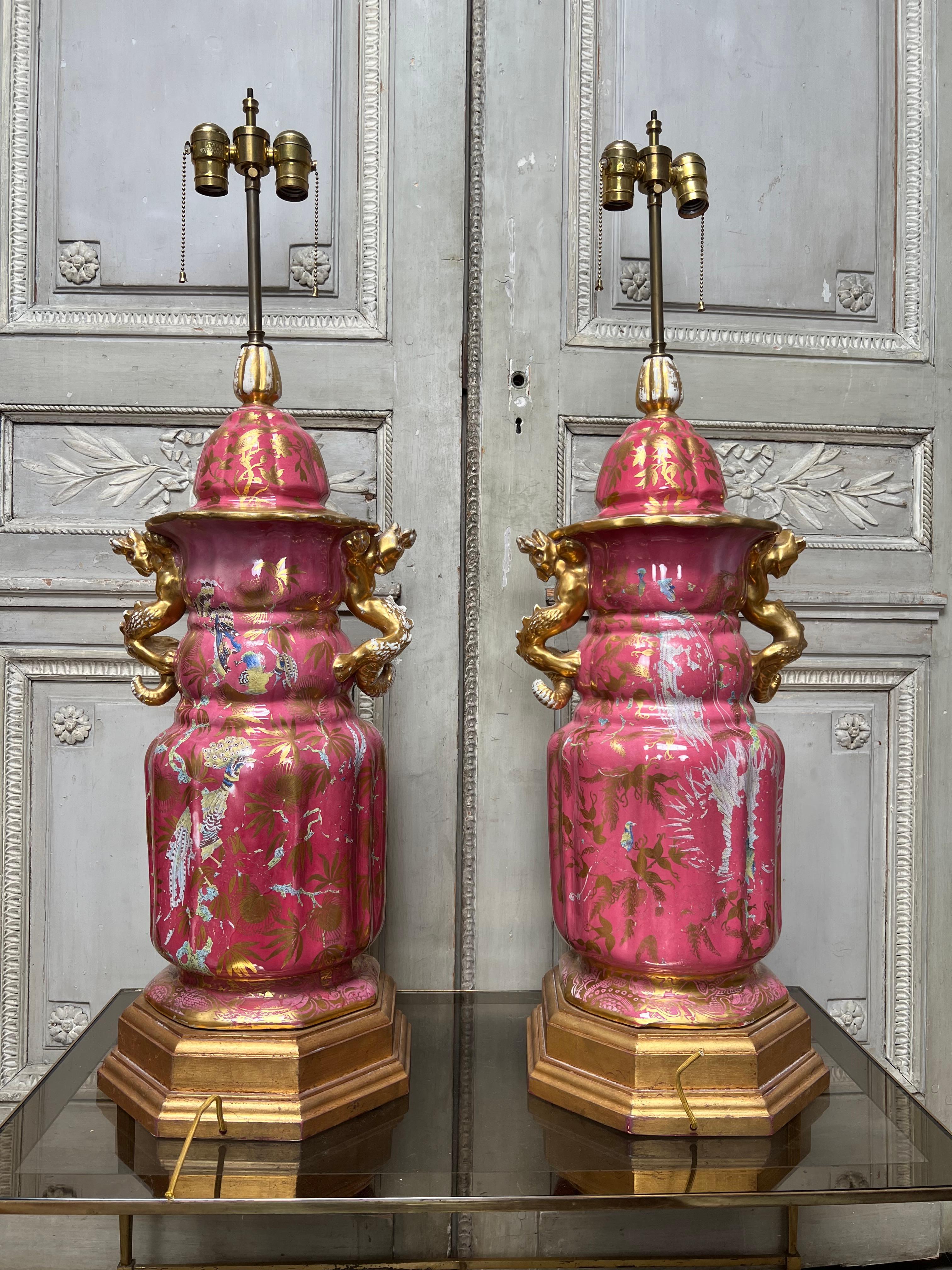 A pair very large English ironstone urns with lids that have been converted to lamp bases.  These pink glazed urns have a an overall gilded decoration as well as polychrome painted birds on the other side, but there is a lot of enamel loss to those