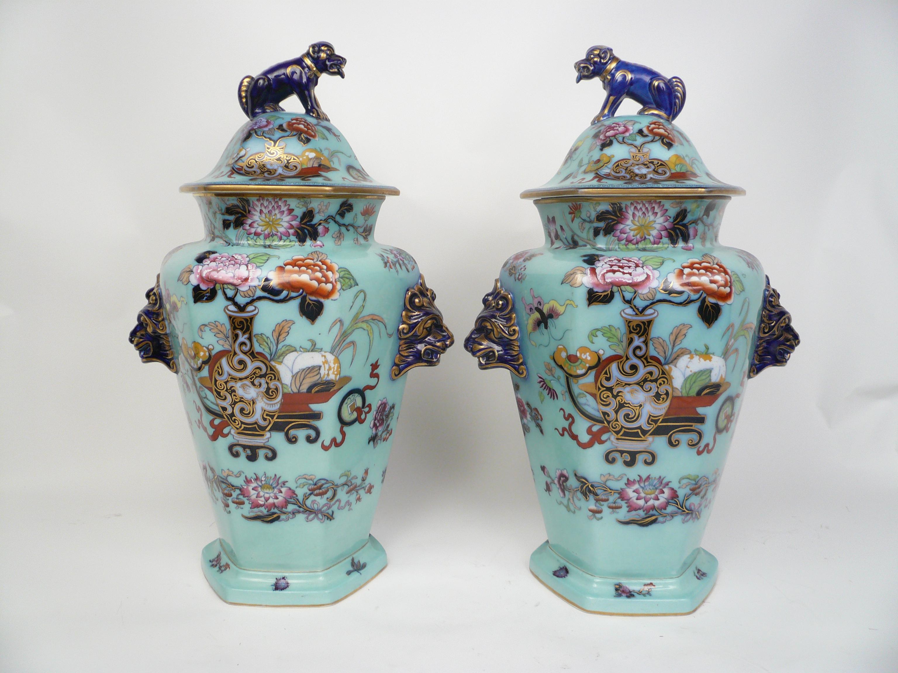 This impressive pair of hexagonal urns and covers are hand painted in the Imari palette, and feature floral decoration. Each vase has gilt highlighted vases, peonies, lion head handles, and Foo dog finials.