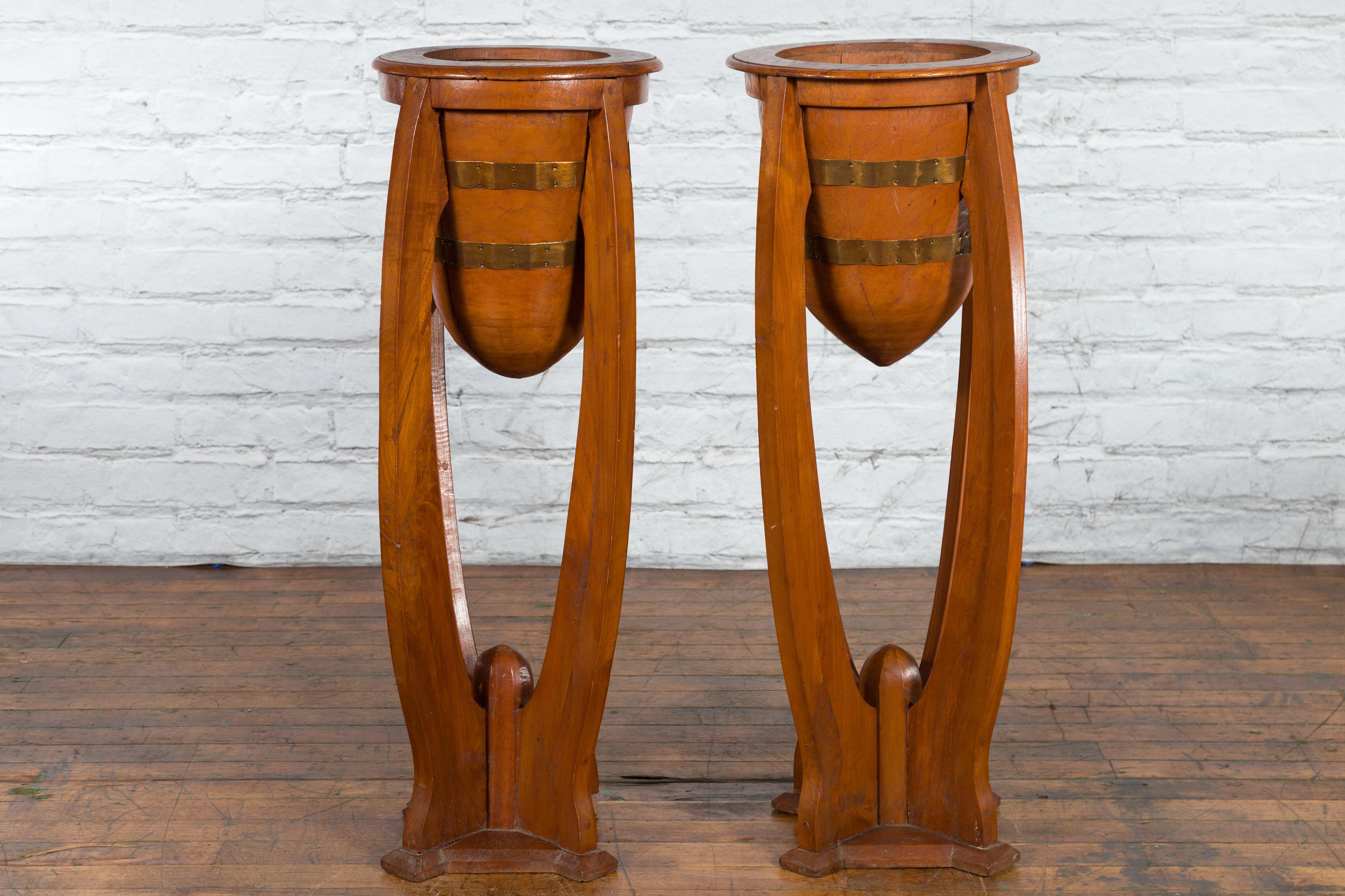 A pair of Javanese Art Deco style teak wood plant stands from the 20th century, with tall proportions, horizontal brass braces and curving bases. Created on the island of Java during the 20th century, this pair of large planters charms us with their