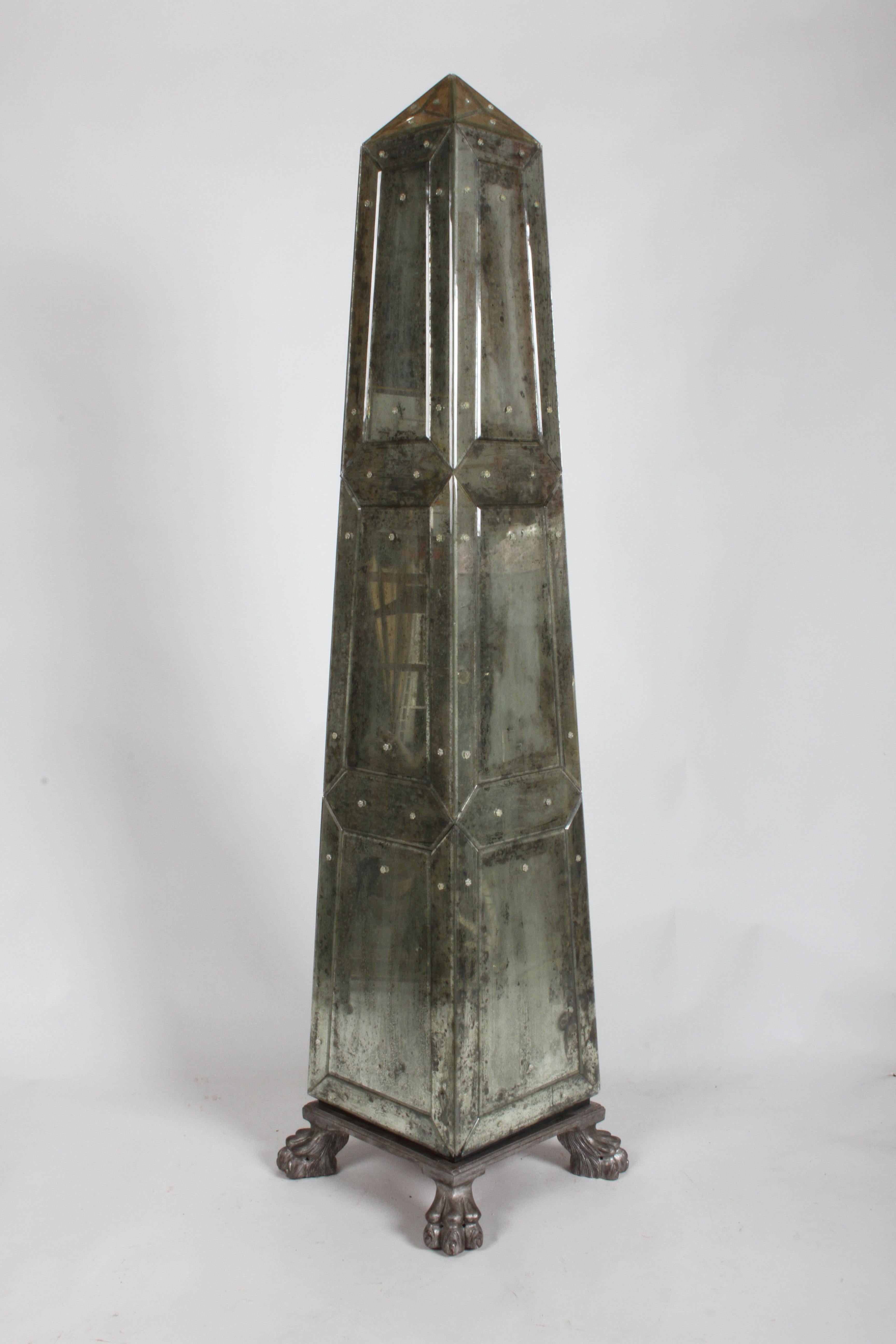Large pair of decorative obelisks in the neoclassical style, possibly Italian. Each fully covered in antiqued or distressed beveled mirrored panels, attached with rosettes. The silver leafed bases have large hairy paw feet and are attached by screws