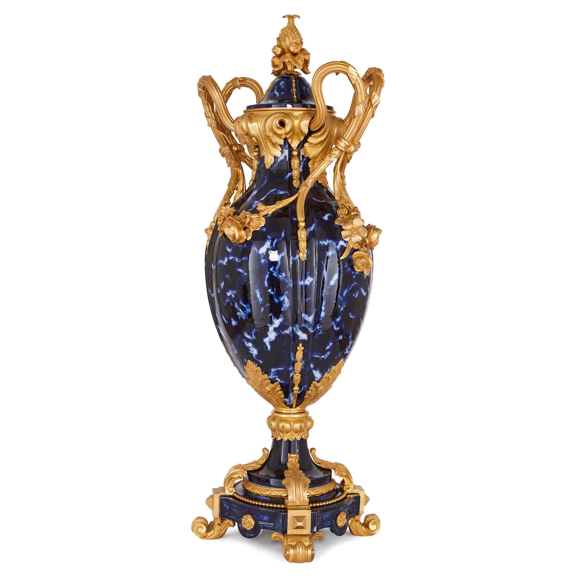 The two vases are of large, ovoid form, and feature fluted bodies which are crafted in ceramic with a mottled blue colouring. The vases are mounted with twin gilt bronze scrolling handles that are joined by floral swags and garlands, and their