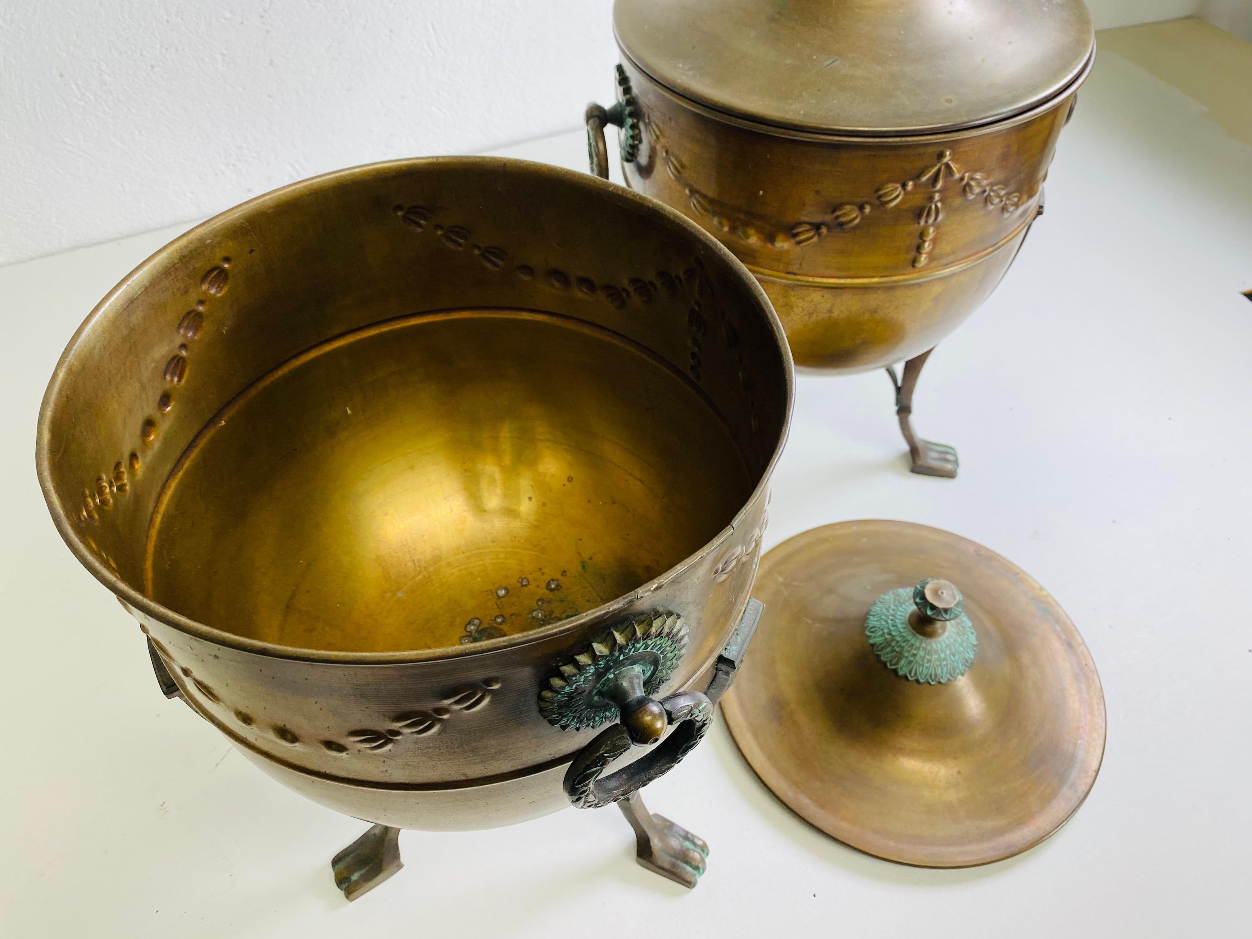 Si tratta di una bellissima coppia di cachepot in rame di ispirazione Regency con coperchio. Questa coppia di pentole in rame ha manici ben dettagliati su entrambi i lati e si regge su tre gambe ben dettagliate. I vasi presentano una delicata