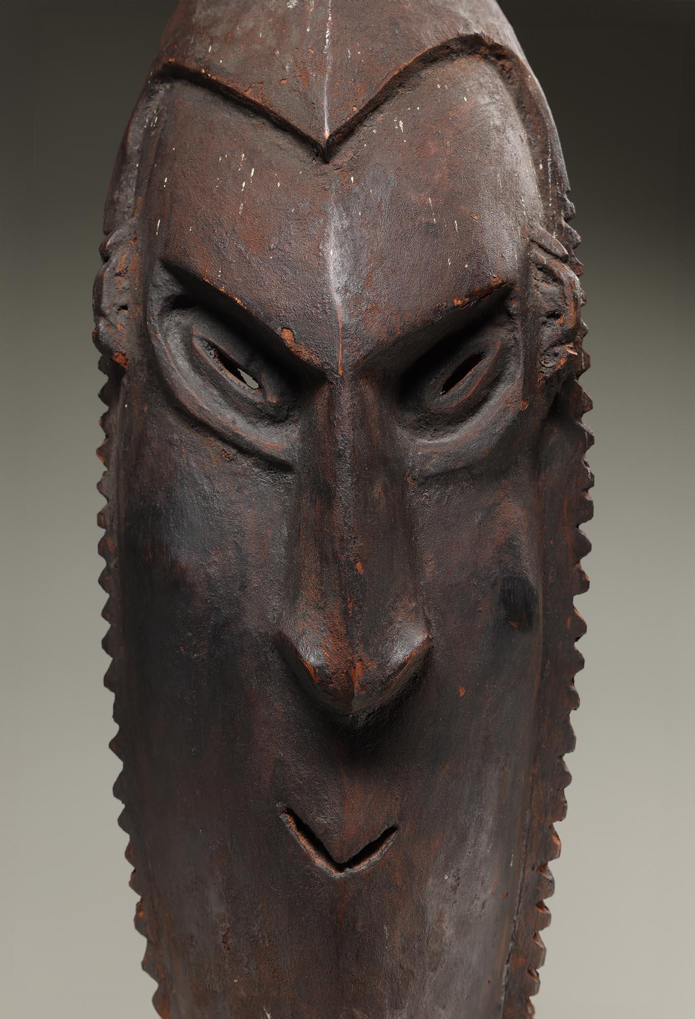 Gran máscara de madera tallada de color marrón rojizo oscuro con restos subyacentes de pigmento rojo, procedente de la zona del río Sepik de Nueva Guinea.  Expresión fuerte mirando hacia abajo, bordes de flecos tallados.  Signos de desgaste y