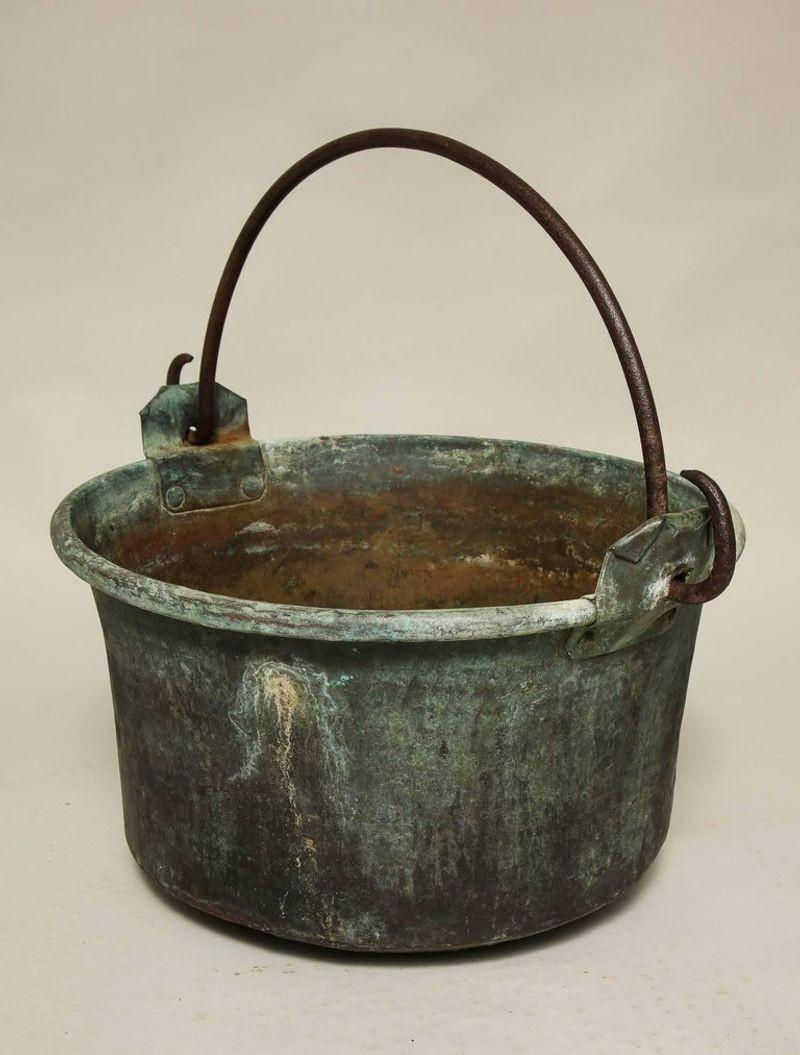 A large copper container with a weathered and patinated surface, flared sides, rolled rim and having a wrought iron carrying handle mounted on riveted hinge ears. Now useful next to a fireplace for logs.