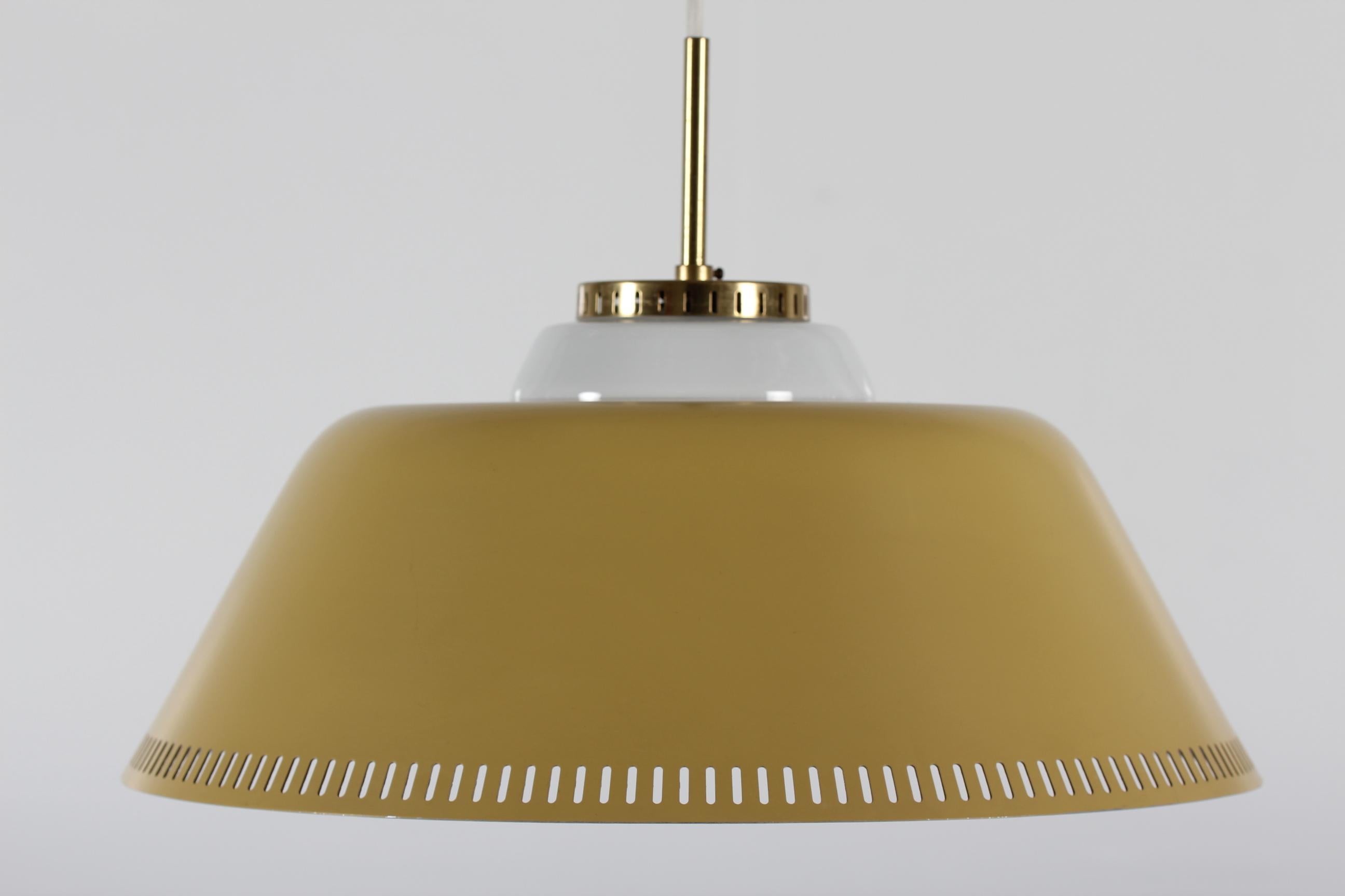 Large modernist pendant light attributed to Bent Karlby and presumably made by Lyfa.
The metal pendant has a dusty yellow lacquer on the outside and the inside is shiny metal. The top is made of brass with lacquer.
A milky white glass inset cover