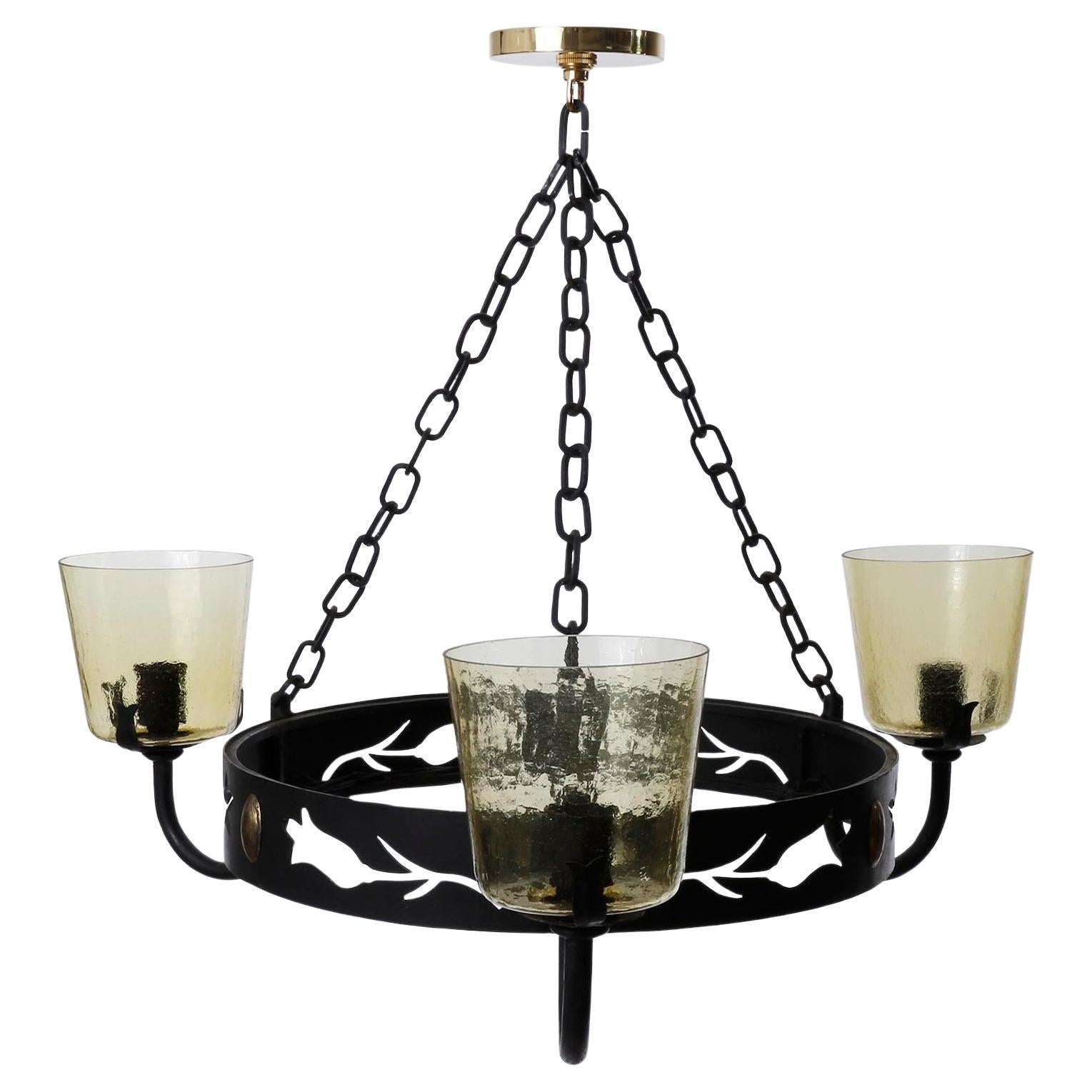 A large three-arm pendant chandelier made of blackened iron and metal with amber tone glass lamp shades manufacturd in Mid-Century, circa 1960 (late 1950s or early 1960s).
The fixture body is made of a round arch of metal and sheet iron with
