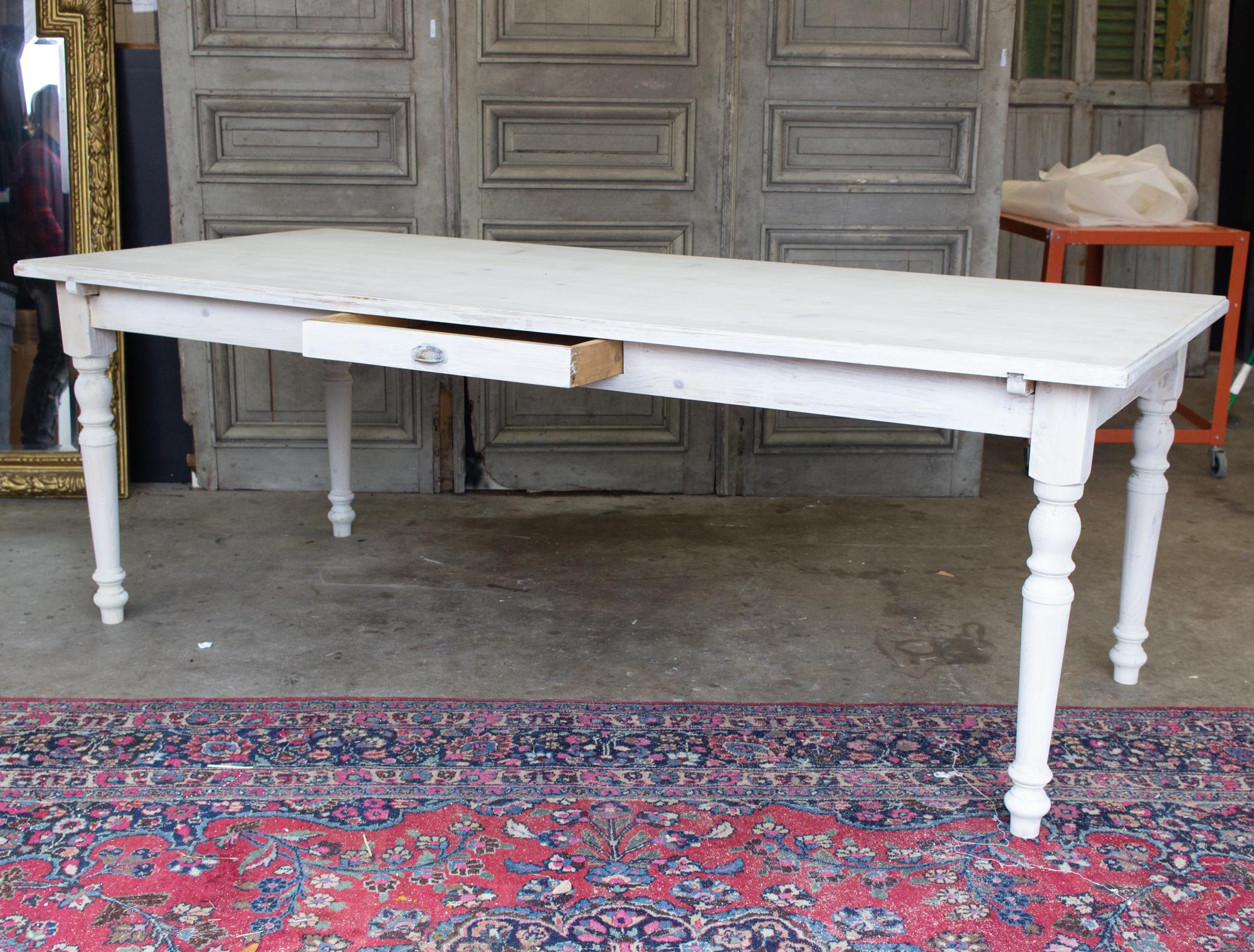 This large, pine farm table was sourced in France and features turned leg details and a single drawer at center. The drawer has a brass cup-pull installed, with some patination to the metal. The whitewash painted finish is new, and still allows the