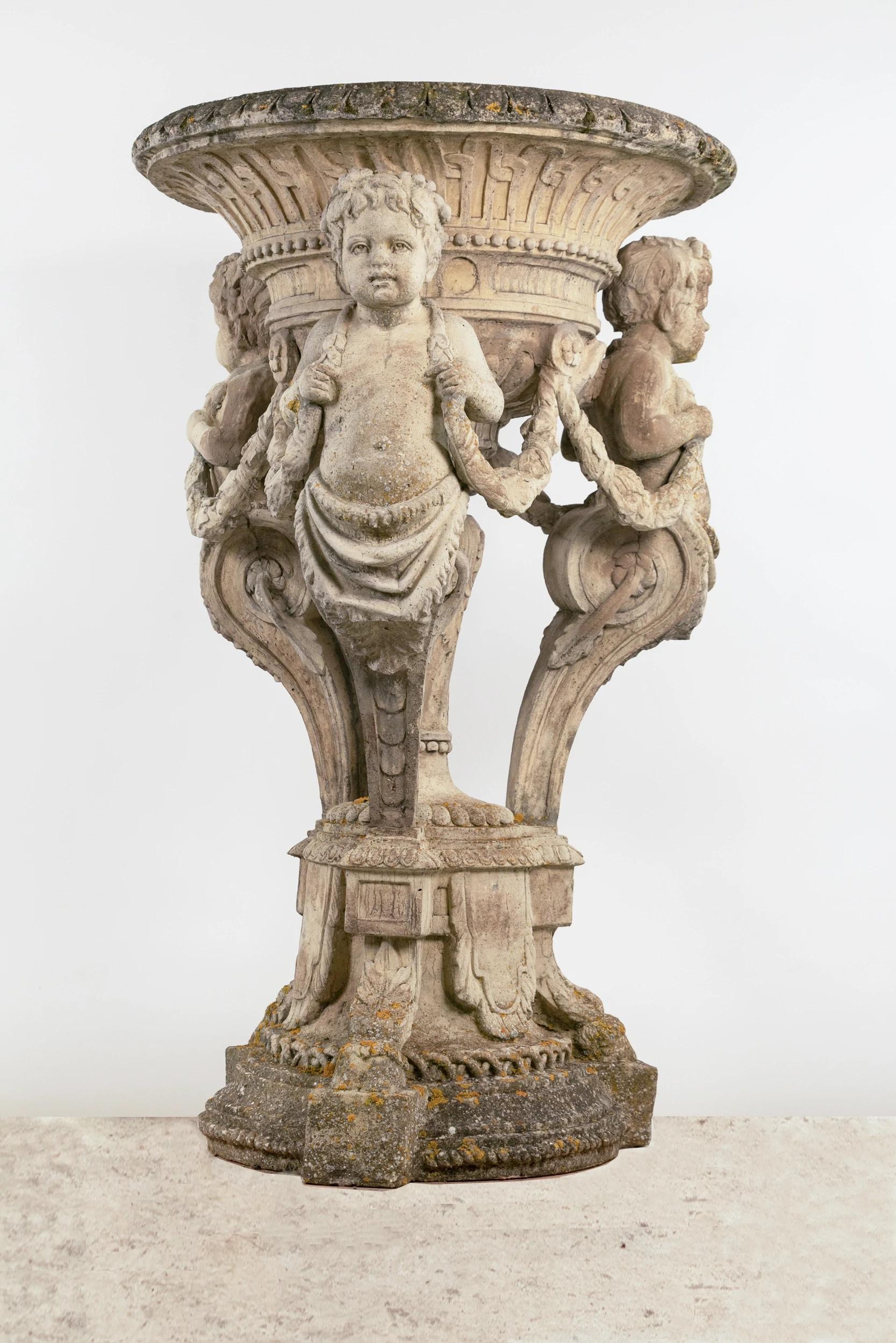 Monumental garden basin in reconstituted stone, depicting three cherubs draped as caryatids carrying a garland of flowers and supporting a large basket decorated with a frieze of pearls, gadroons, cartouches and interlacing. Base also richly