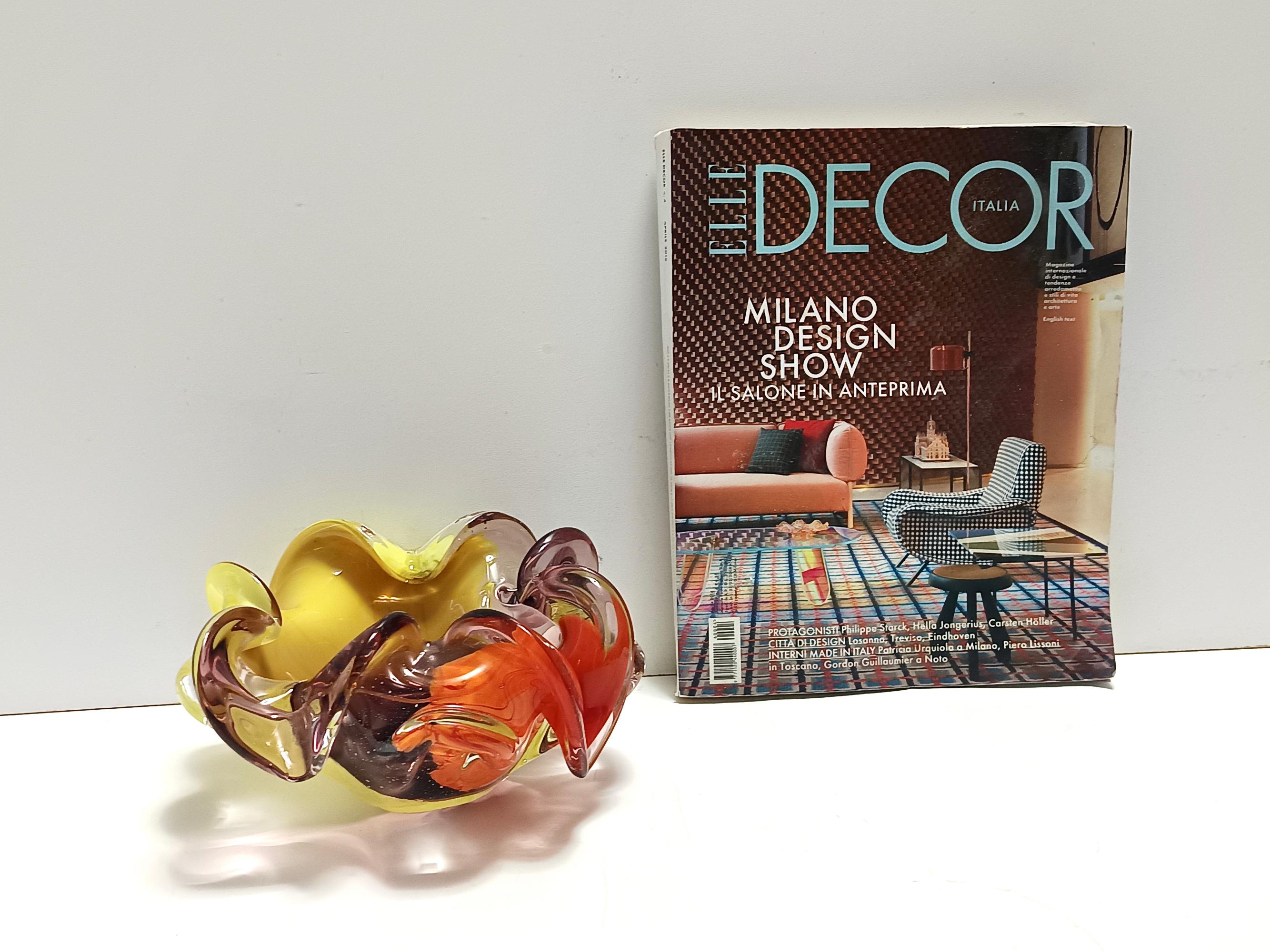 Fabriqué en Italie, années 1960.
Ce cendrier / attrape-tout en forme de fleur est réalisé en verre de Murano jaune, rouge corail et violet clair modelé à la main.
Il contient également de la poussière blanche. 
Il s'agit d'une pièce vintage, qui