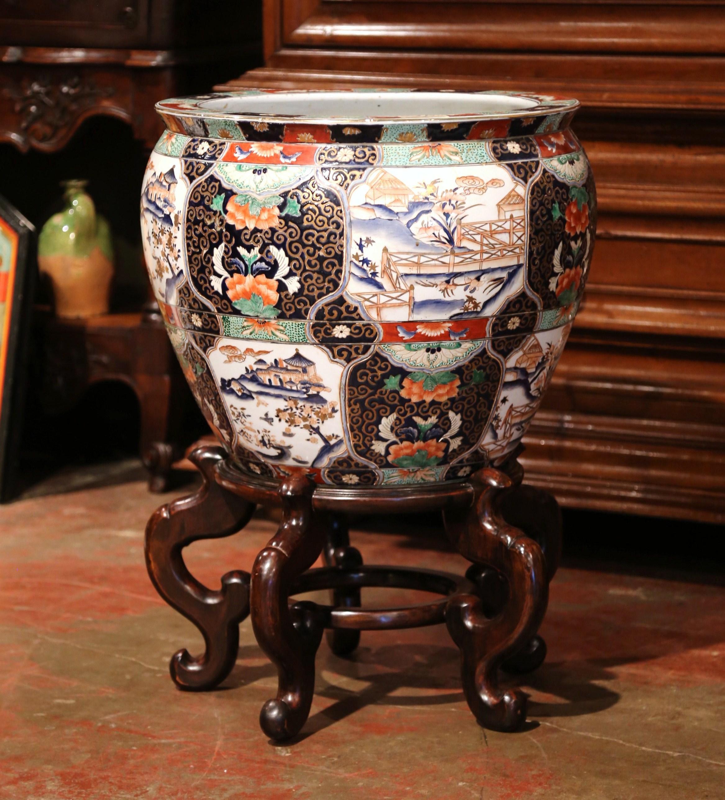This elegant and colorful vintage fishbowl sited on carved fruitwood base was created in China, circa 1960. Round in shape, the large, exotic porcelain planter features Classic oriental floral and landscape scenes throughout. The ceramic bowl is