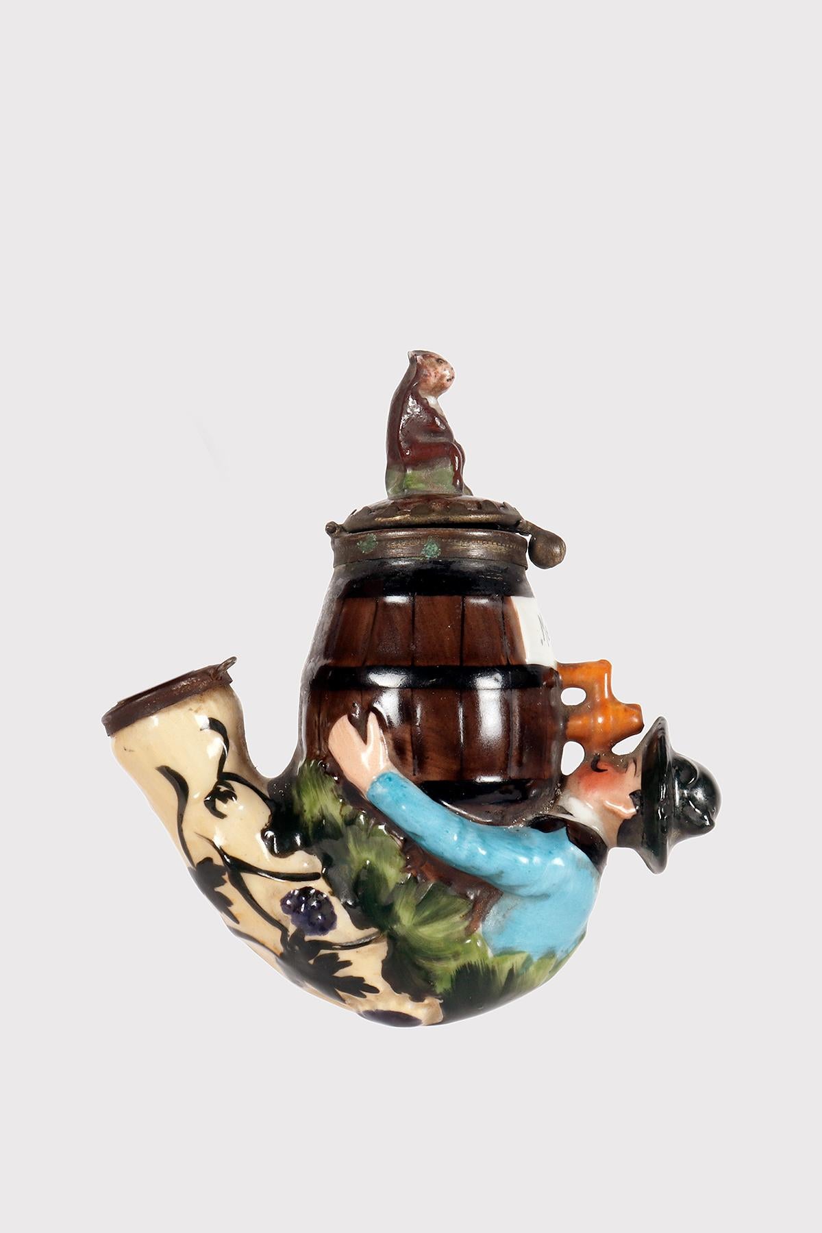 Painted porcelain pipe, with opening bowl, depicting a man with a hat, lying down and embracing a barrel of Alsatian wine from Metz, with his mouth attached to the tap from which the wine comes out. On the lid, a monkey sits, looking at it. The long