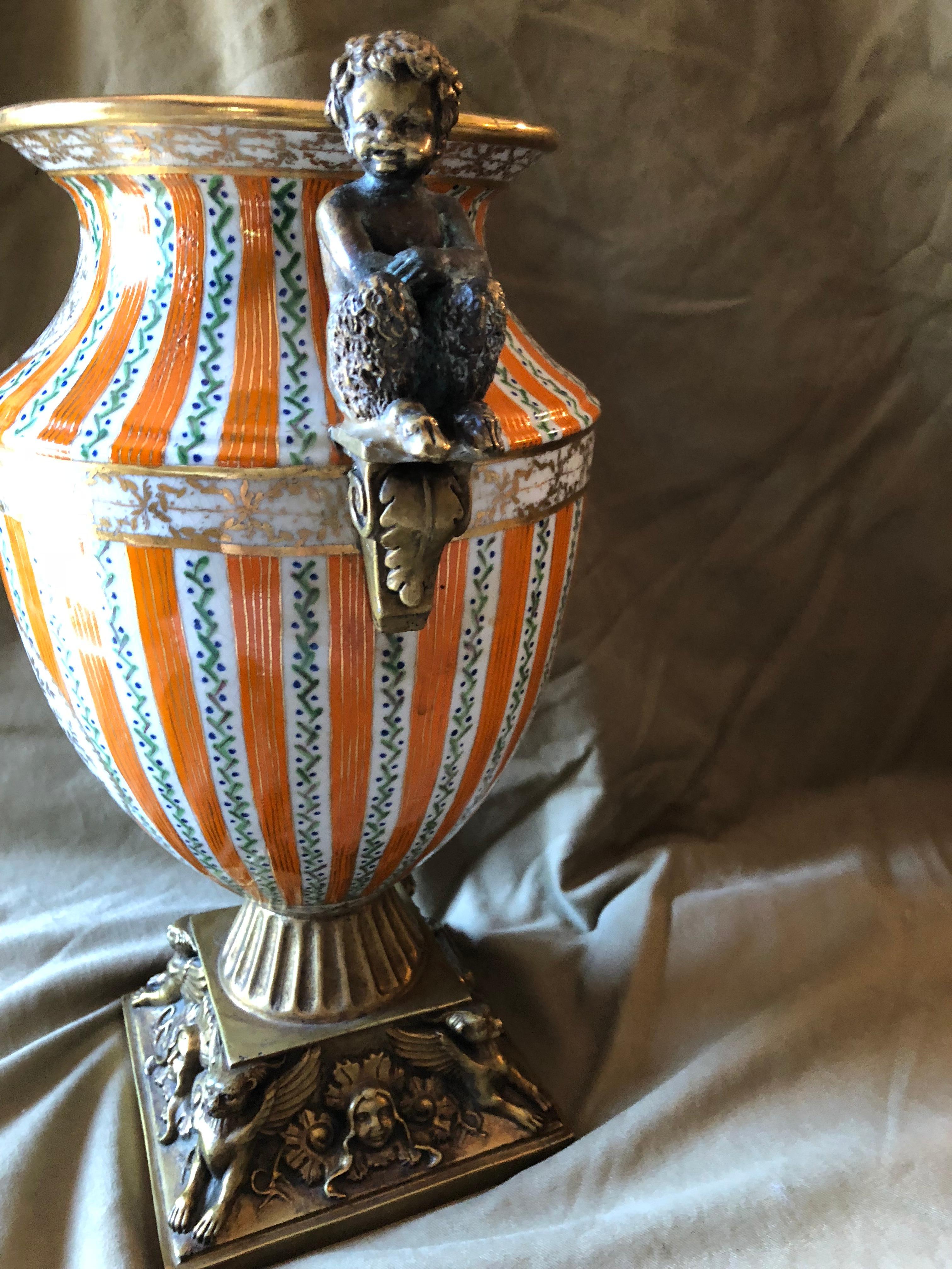 This is a good sized heavy porcelain urn, beautifully decorated in orange and white stripe and gold. Its brass handles are of fauns sitting on a bracket. The base is also decorated brass.