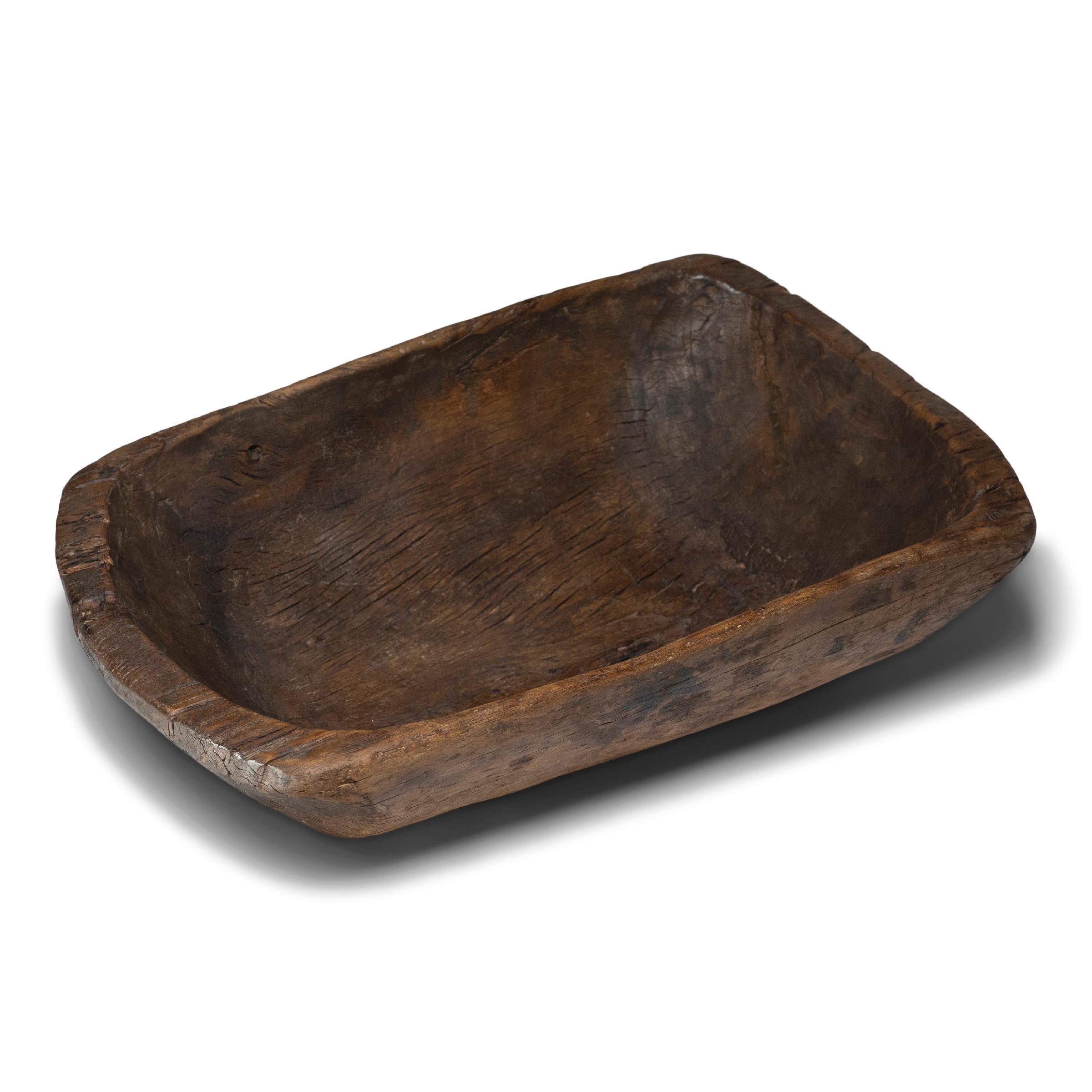 This large wooden farm tray from perfectly balances rustic texture with minimalist simplicity. Carved from a solid block of wood, the tray has a rectangular shape with rounded corners, sloping sides, and a deep interior basin. Years of use have