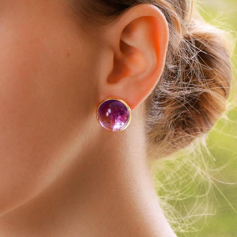 A vibrant pair of purple amethyst earrings set in 18k yellow gold.

Each earring is predominantly set with a large cabochon amethyst stone, bezel set in a simple yet elegant setting. The earrings are secured to reverse with a post and clip