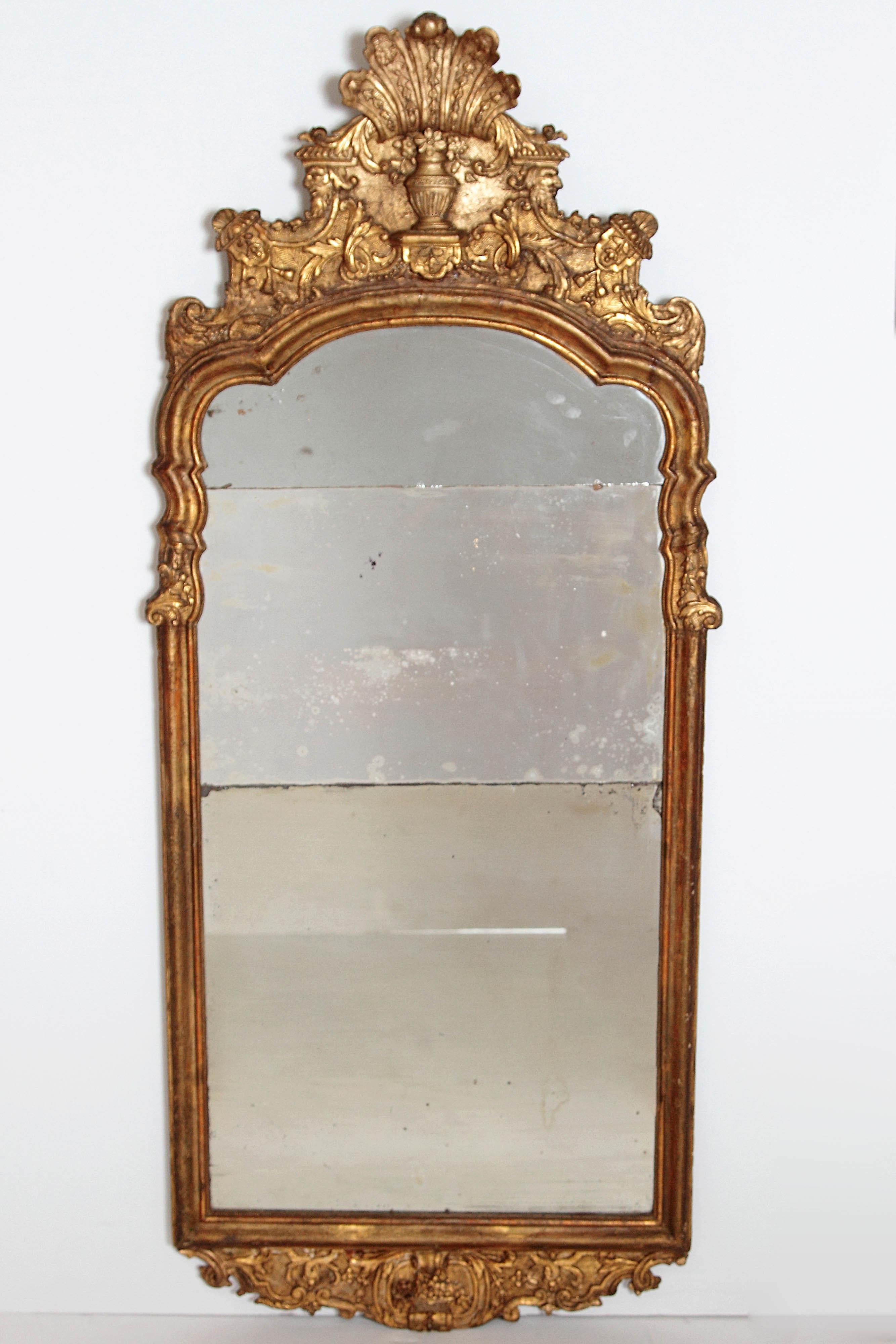 A large period Queen Anne gilt-gesso mirror in gilt frame with elaborate crown and base. Intricate crosshatching in background with acanthus leaf and foliage carvings. Crown has urn with five feather forms at top. Mirror plate is in three parts.