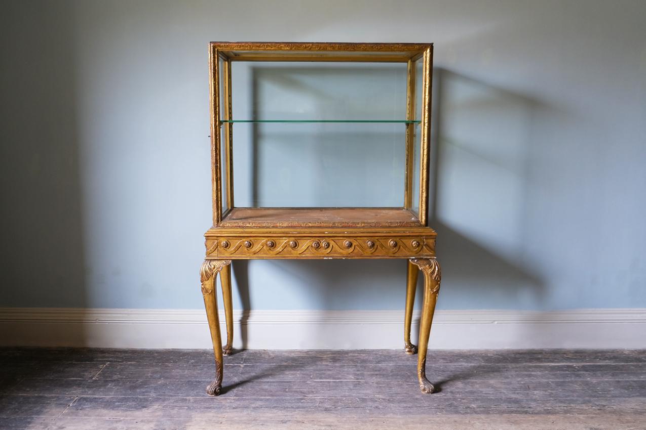 A 20th century Queen Anne style gilt-gesso display cabinet

The display cabinet is c. 20th century, with foliate carvings and intricate guilloche mouldings present throughout. The rectangular top sits above an enclosing drawer, the interior is lined
