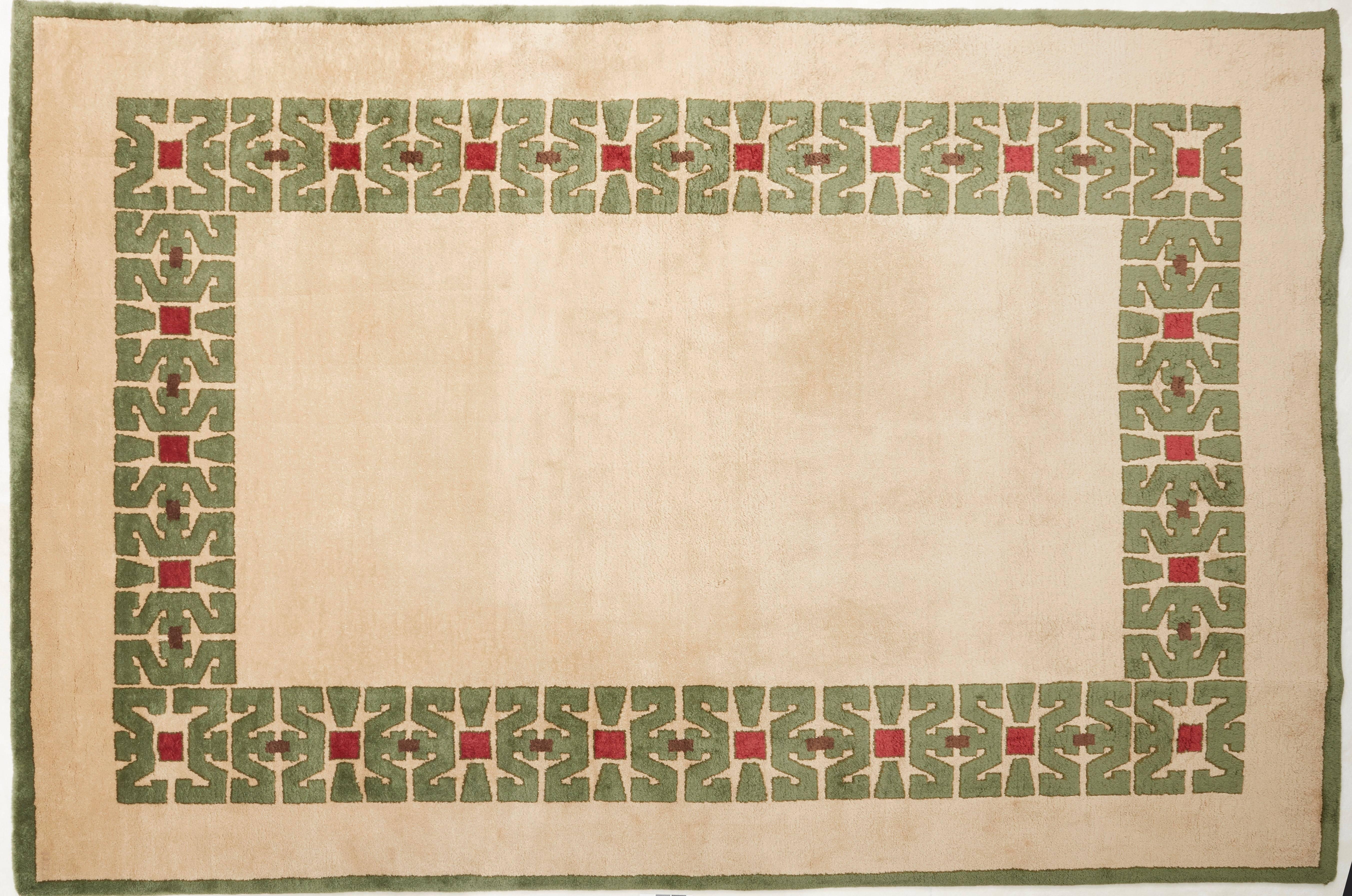 A Large knotted woollen rug with beige ground and green border, central decoration forming a frame with green, red and brown Aztec designs.