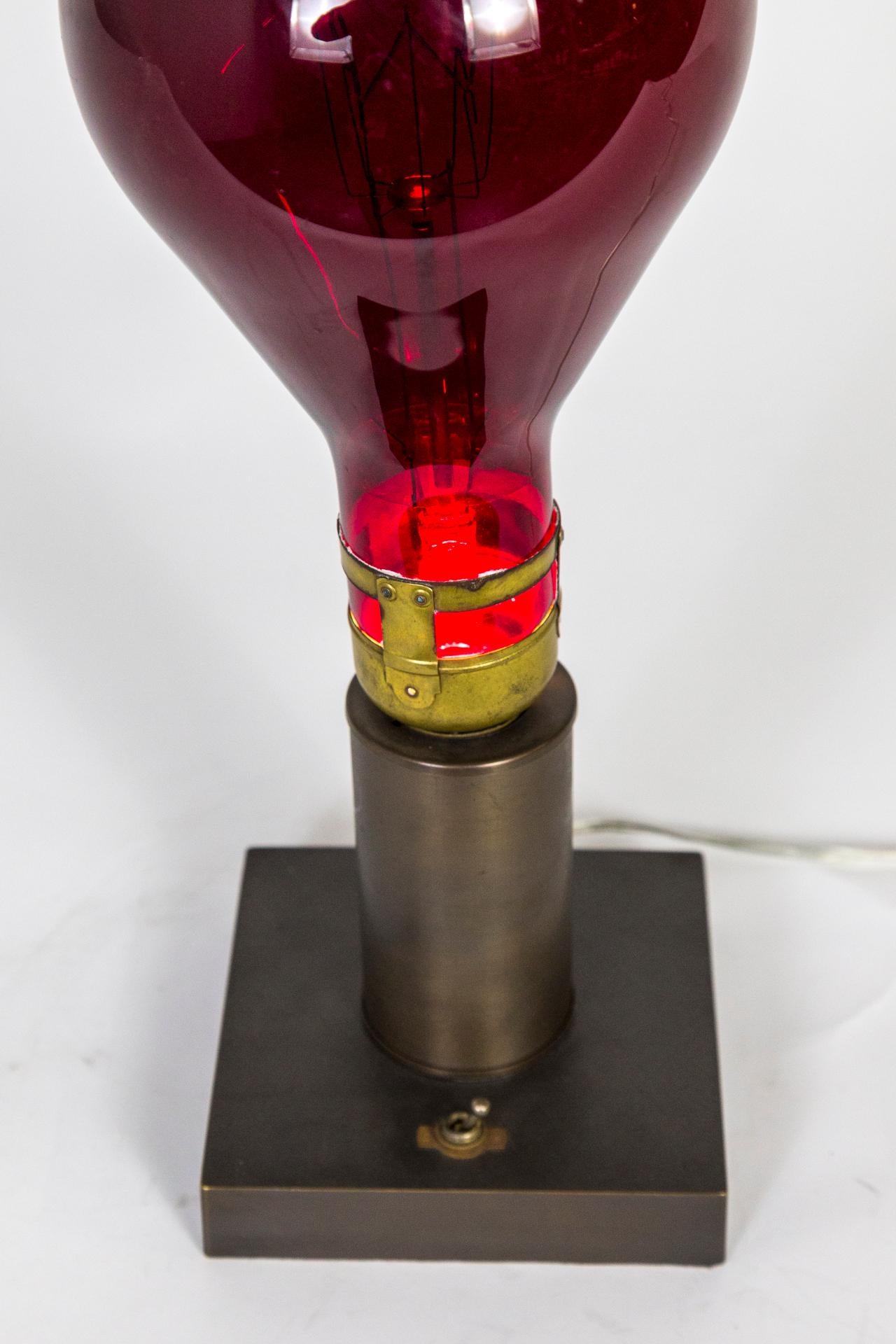 Une lampe de table fabriquée à partir d'une ampoule vintage géante de couleur rouge, éclairée de l'intérieur par une LED e12 Emery Allen (5 watts). Finition soignée avec un interrupteur vintage et une base carrée en laiton teinté dans un ton