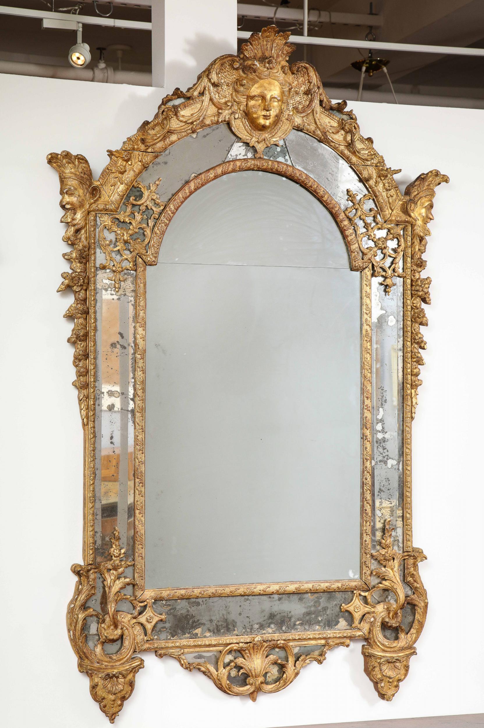Large and fine French Régence period giltwood mirror, the lobed crest with maiden's head mask flanked by foliate and scroll carving, the sides with satyr heads wearing feather headdresses, having foliate carved festoons, the central plates with