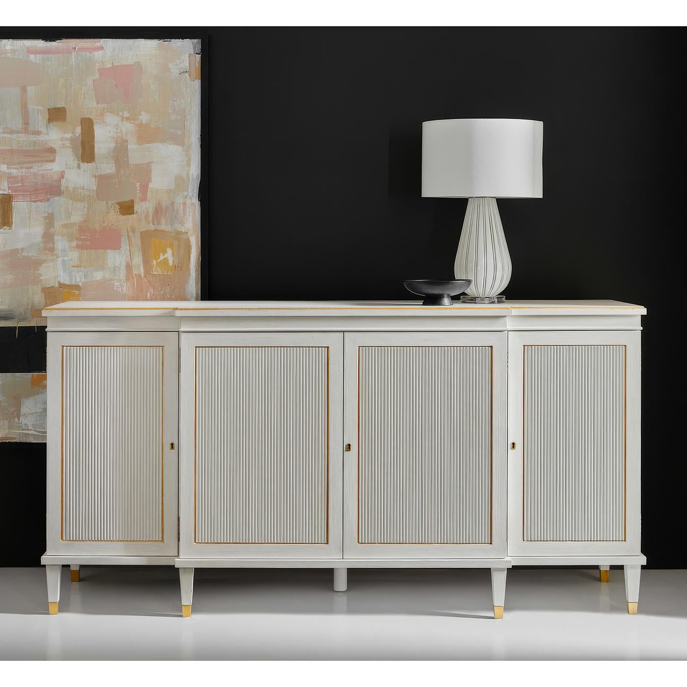 Credenza combines sophisticated style with delicate gilt accents that highlight its classical architectural details. The antique white painted finish provides an elegant backdrop for the reeded door panels, while antiqued gold leaf trim frames each
