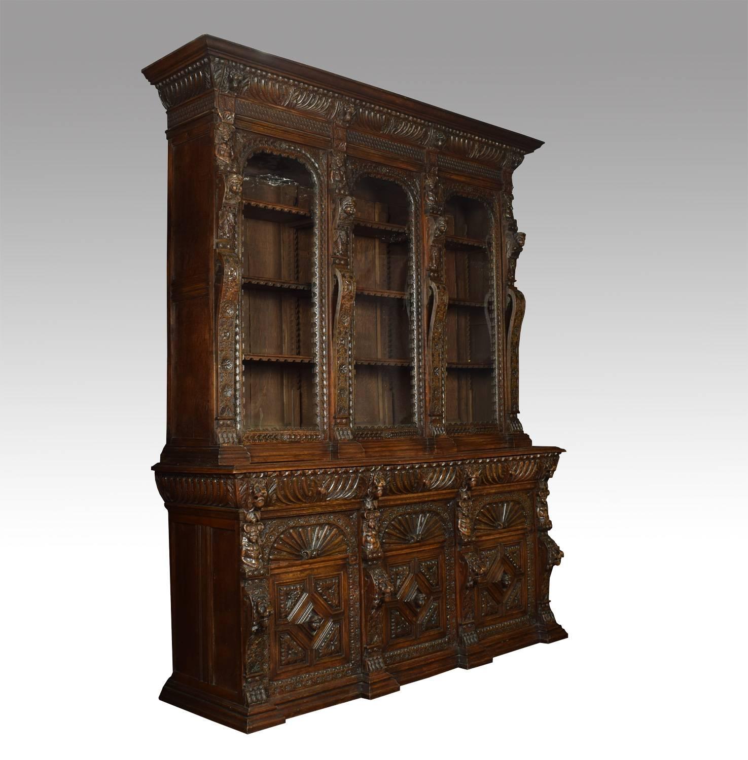 Large Renaissance Revival carved oak three-door bookcase, the deep cornice above a chip-carved and gadrooned frieze above three large glazed doors enclosing adjustable shelved interior, flanked by pilasters carved with lion and figural masks; the