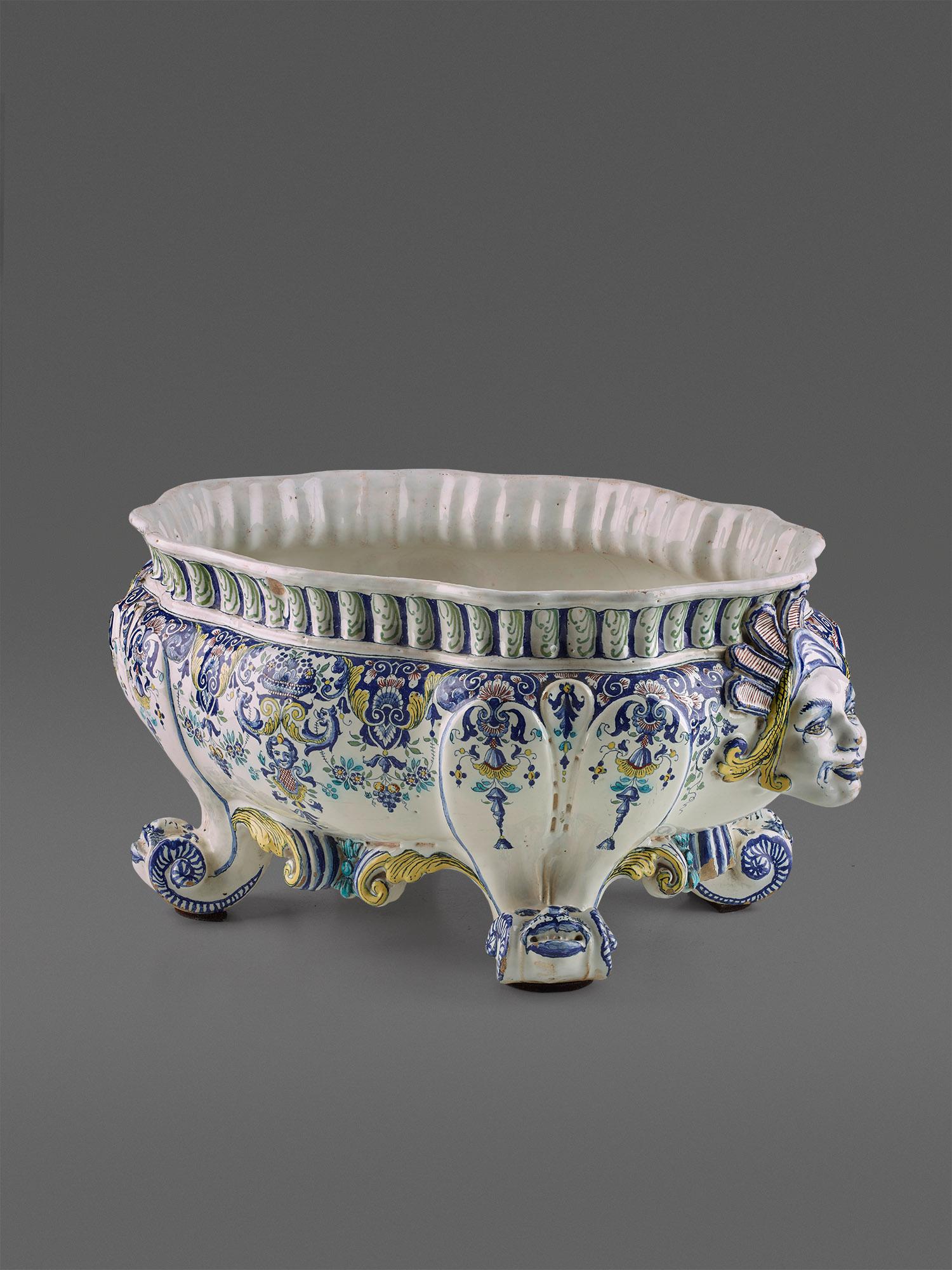 Faience, white glaze with sharp fire decoration in yellow, green and blue tones in delicate gradations. Flared volute feet with dolphins support the oval
support the oval, passively curved planter with fully sculptured female busts as decorative