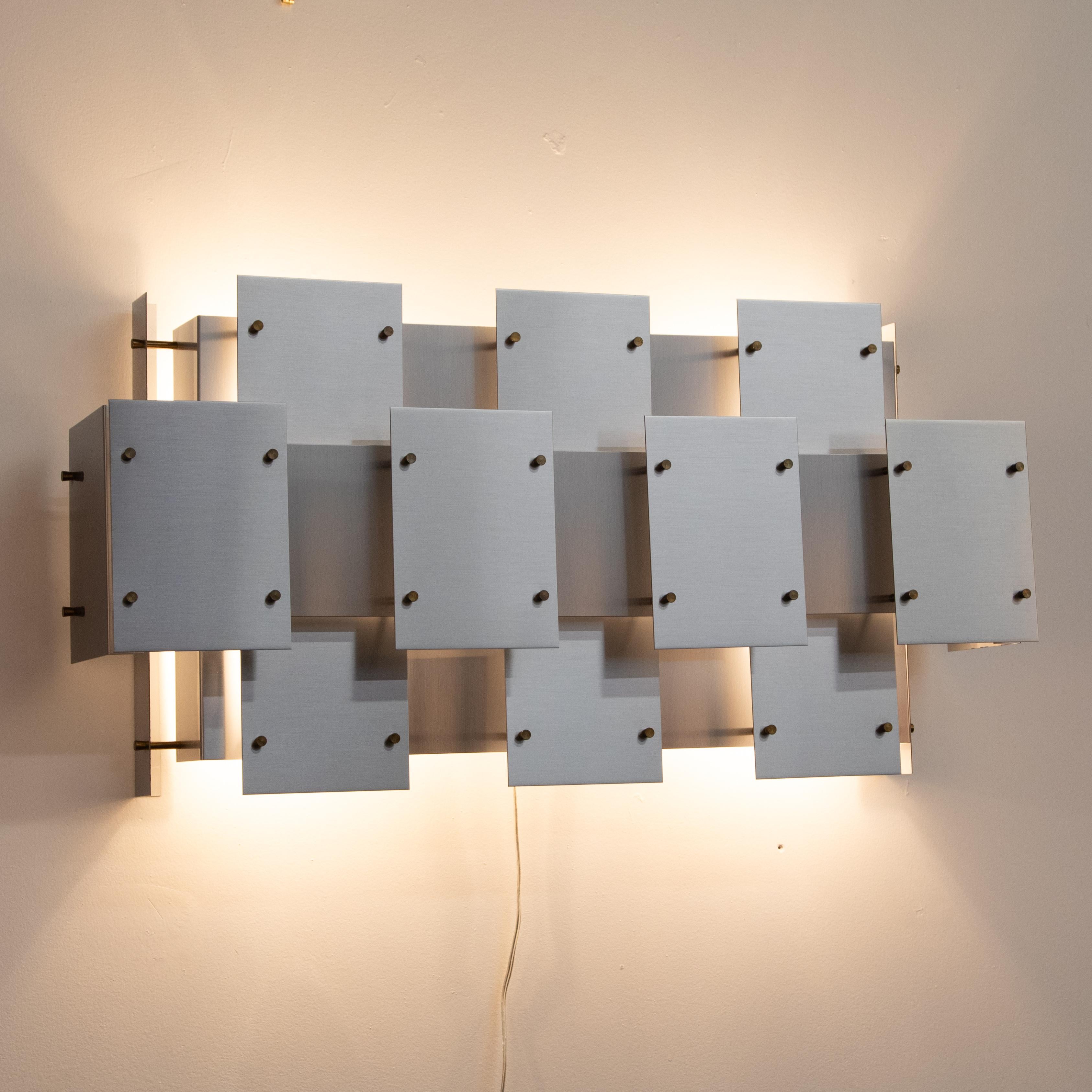 This interlocking geometric wall sconce features brushed-steel tiles secured with brass fittings. The layers of tiles cover the bulbs completely, and spacing between the tiles allows for soft, indirect light. Porcelain sockets hold four