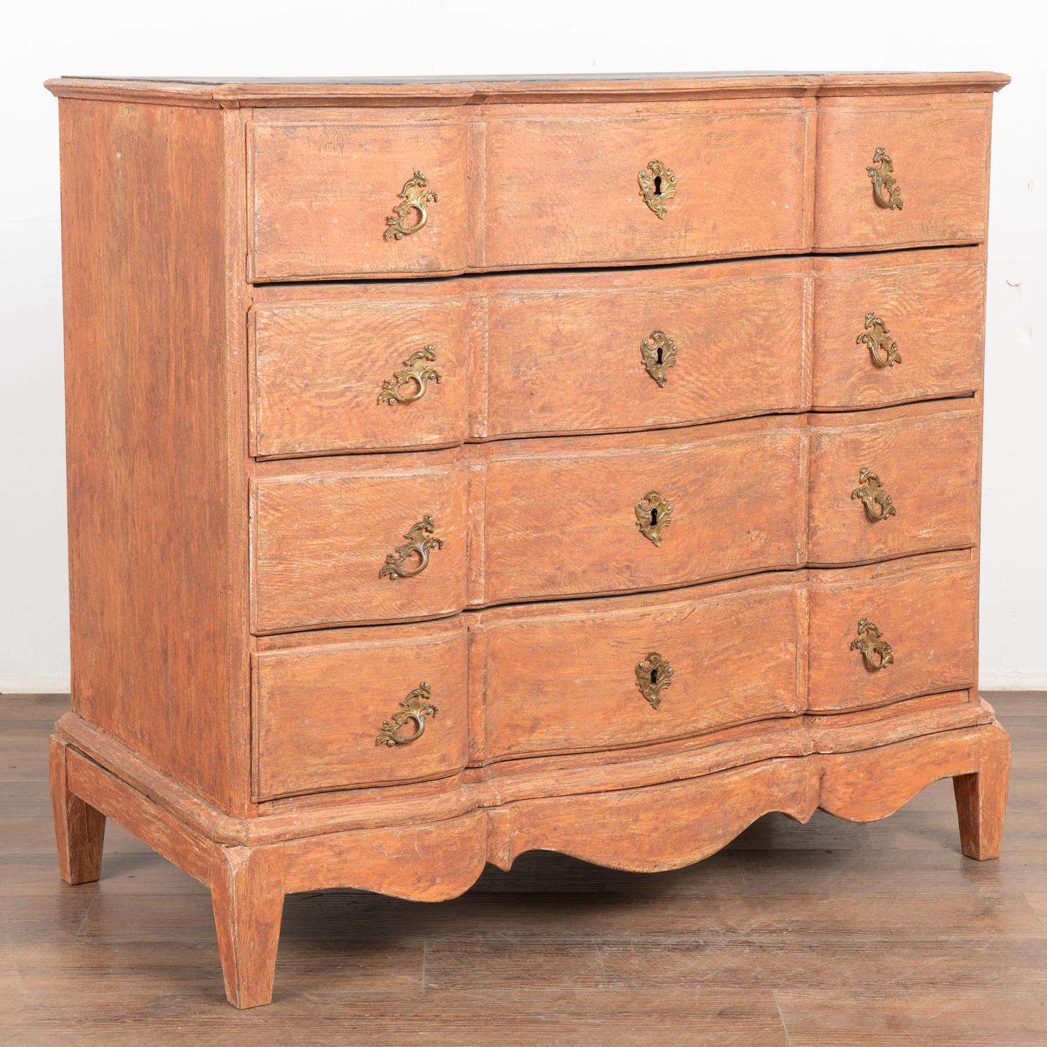 This antique rococo large oak chest of drawers features a serpentine front, brass hardware pulls and is raised on a scalloped skirt base.
The newer, professionally applied and layered salmon painted finish has pink undertones and is gently scraped