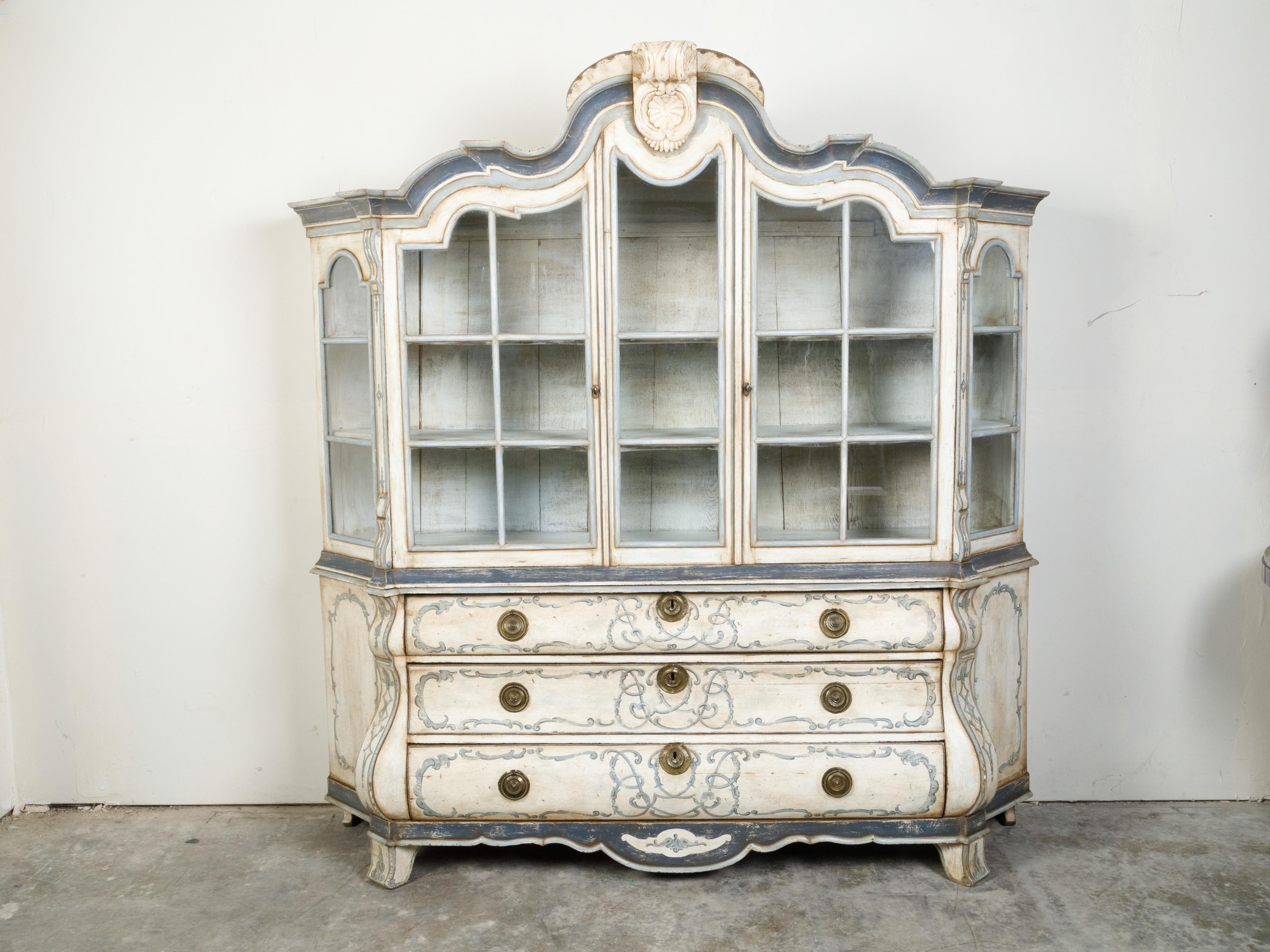 A Dutch painted and carved wooden cabinet from the 19th century, with glass doors and bombé chest. Created in the Netherlands during the 19th century, this large Rococo style cabinet features a carved crest sitting above two glass doors with arched