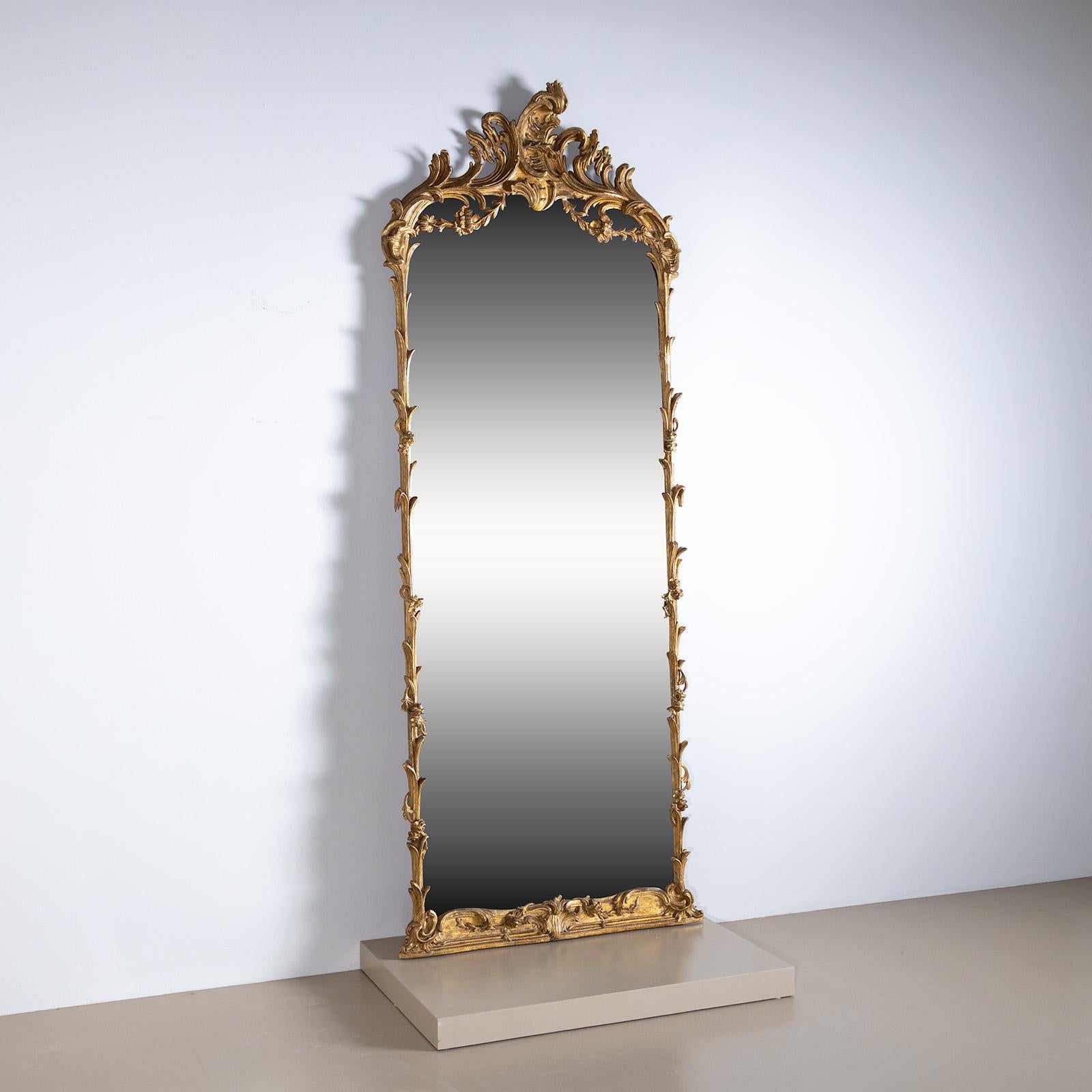 Grand miroir mural dans un cadre en bois et stuc doré, conçu dans un style végétal. Le cimier asymétrique est richement orné de motifs de rocaille et de festons. La partie inférieure, plus prononcée, présente des motifs décoratifs en forme de