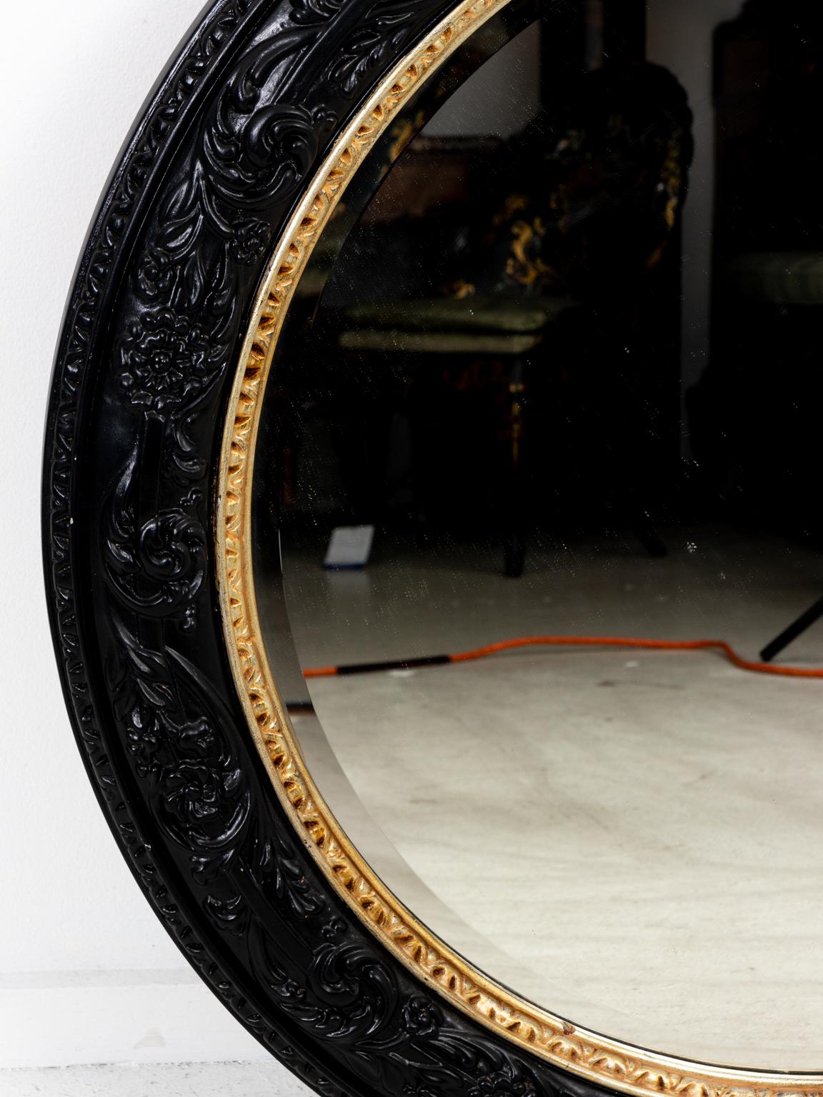 Large round black and gold mirror in the Hollywood Regency style with raised floral and vine motif surrounded by outer egg-and-dart molding with interior gilded egg-and-dart molding, circa 1980s. The mirror also features a beveled edge. Made in the