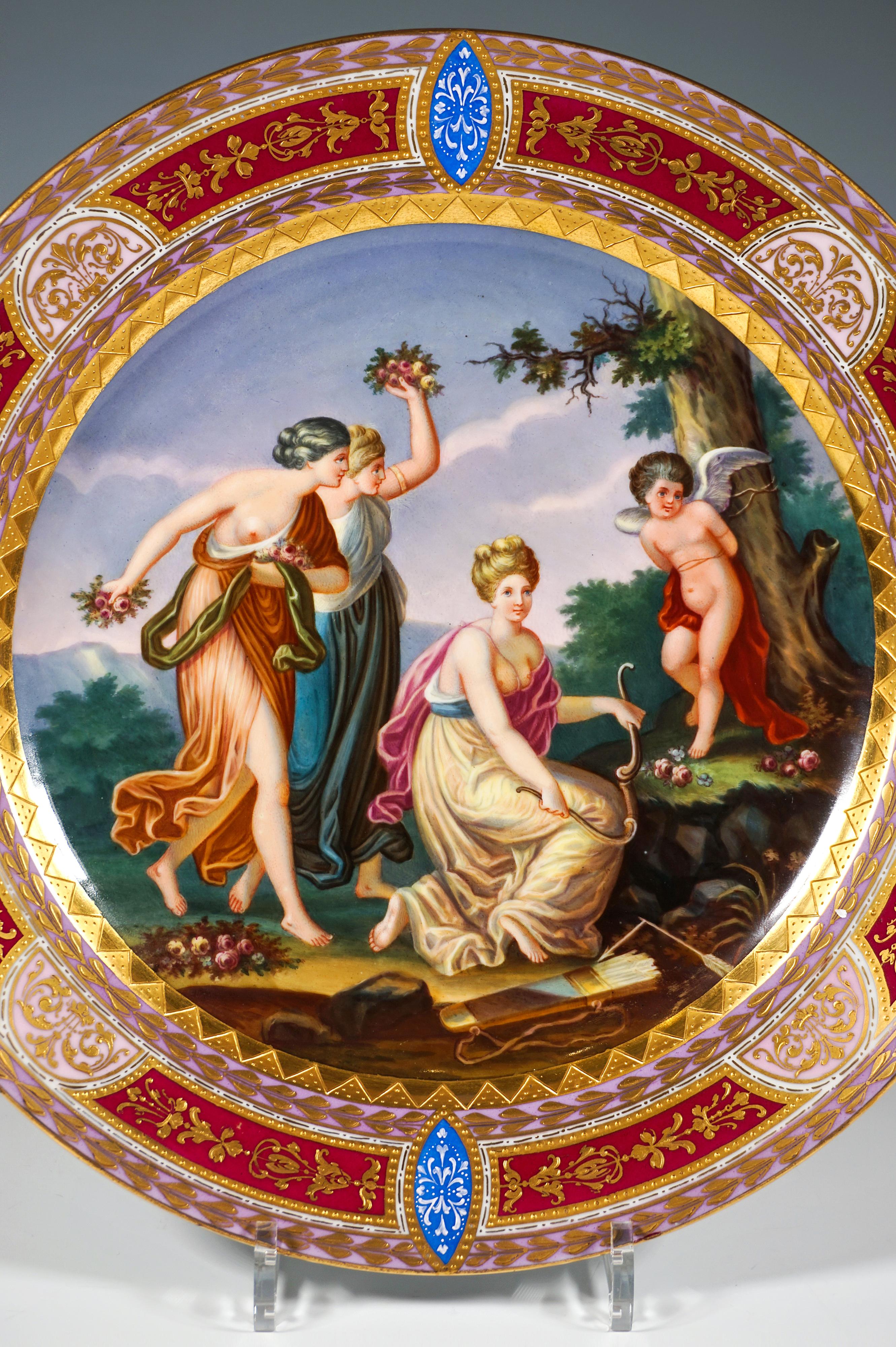 Grande assiette en porcelaine au décor très élaboré : le centre est entièrement occupé par une image polychrome finement peinte, encadrée par une large bande dentelée peinte en or sur or : La scène montre une dispute entre les trois grâces et