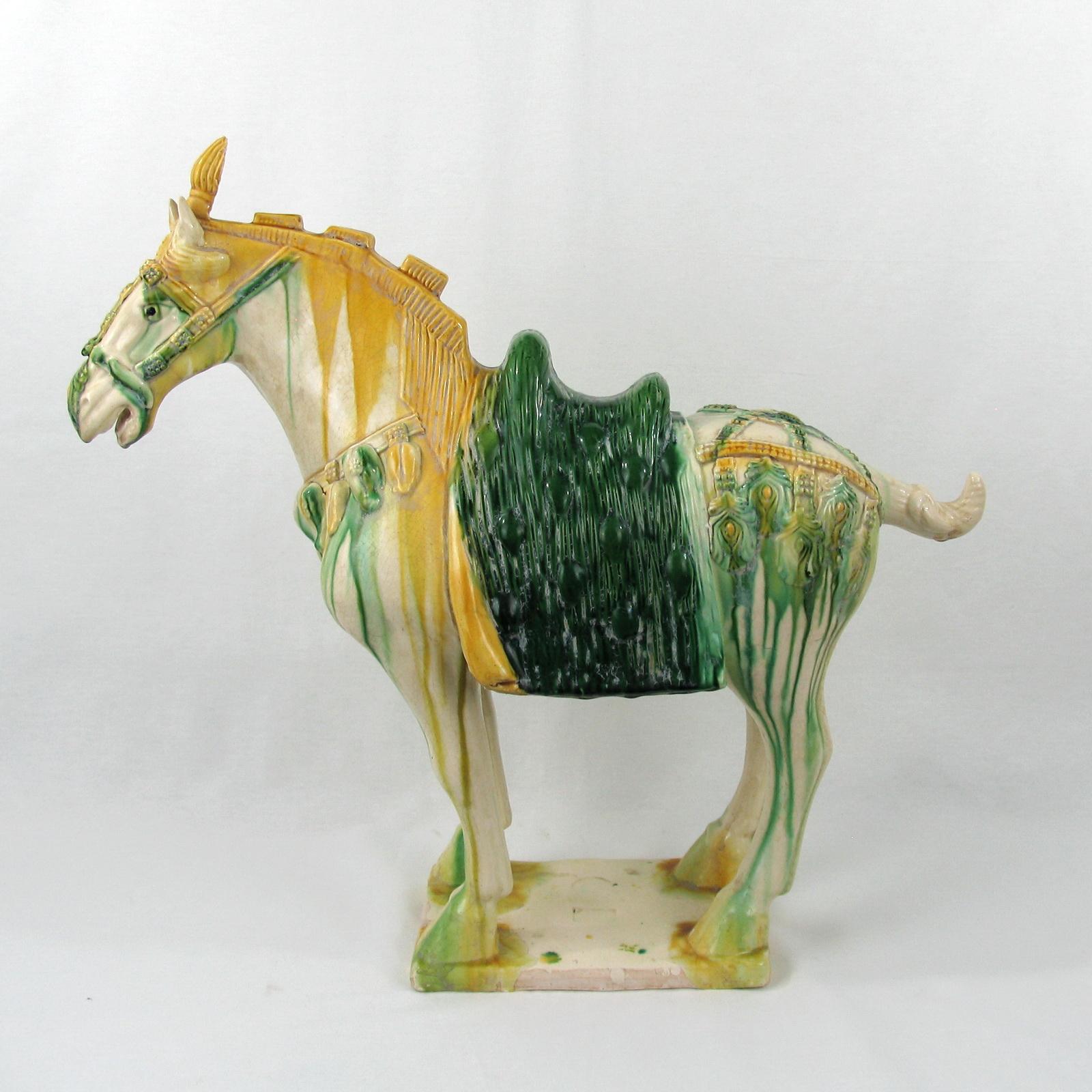 Chinese Tang manner Sancai-glazed pottery statue of a horse standing on all four legs atop a rectangular base. The horse is decorated with bells and flowers. A traditional green saddle completes the look. Illegible impressed marks to the base, rest