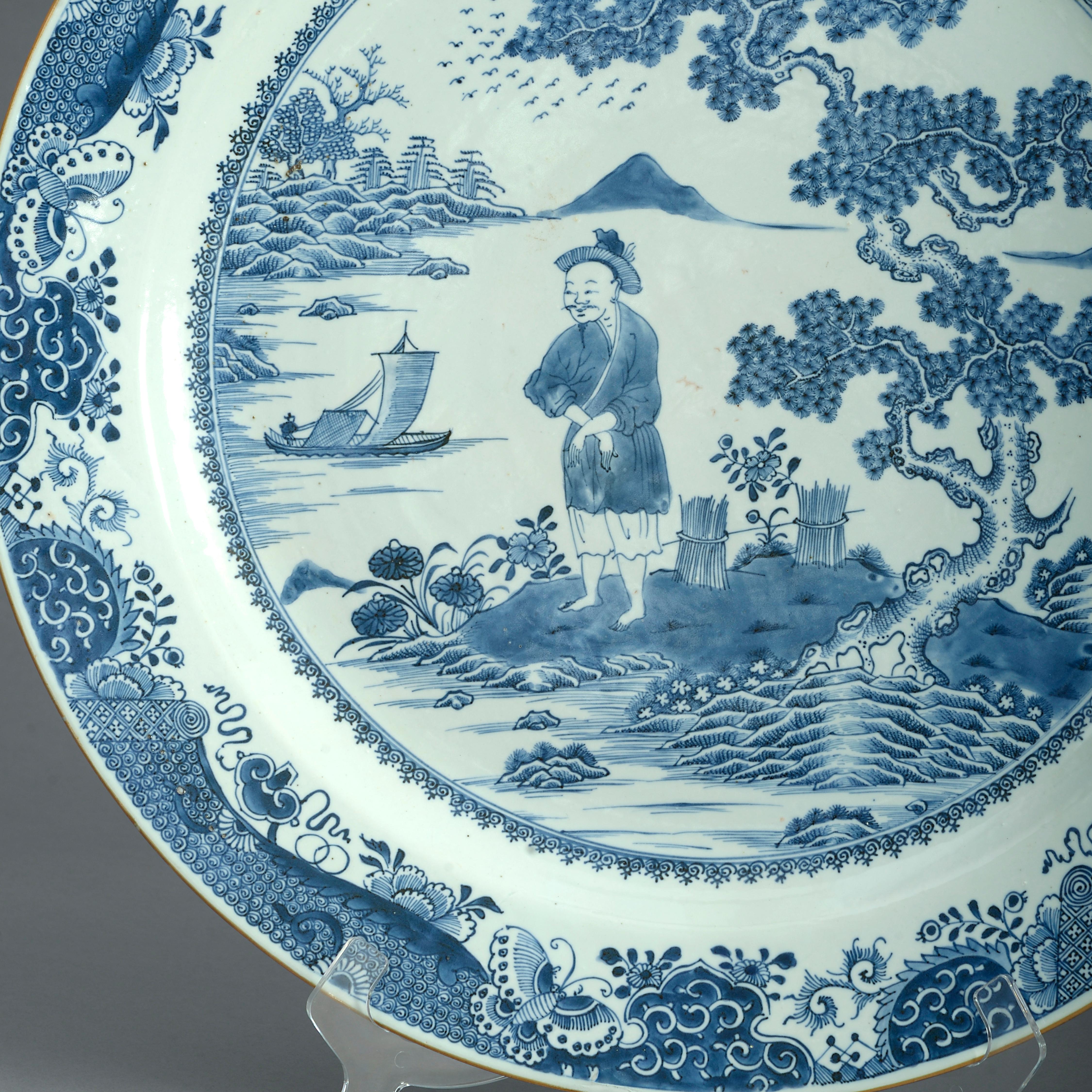 A mid-eighteenth century blue and white porcelain charger, the central panel decorated with a farmer harvesting wheat upon an island within a watery landscape depicting a fishing boat before mountains and hills, all set within a border of auspicious