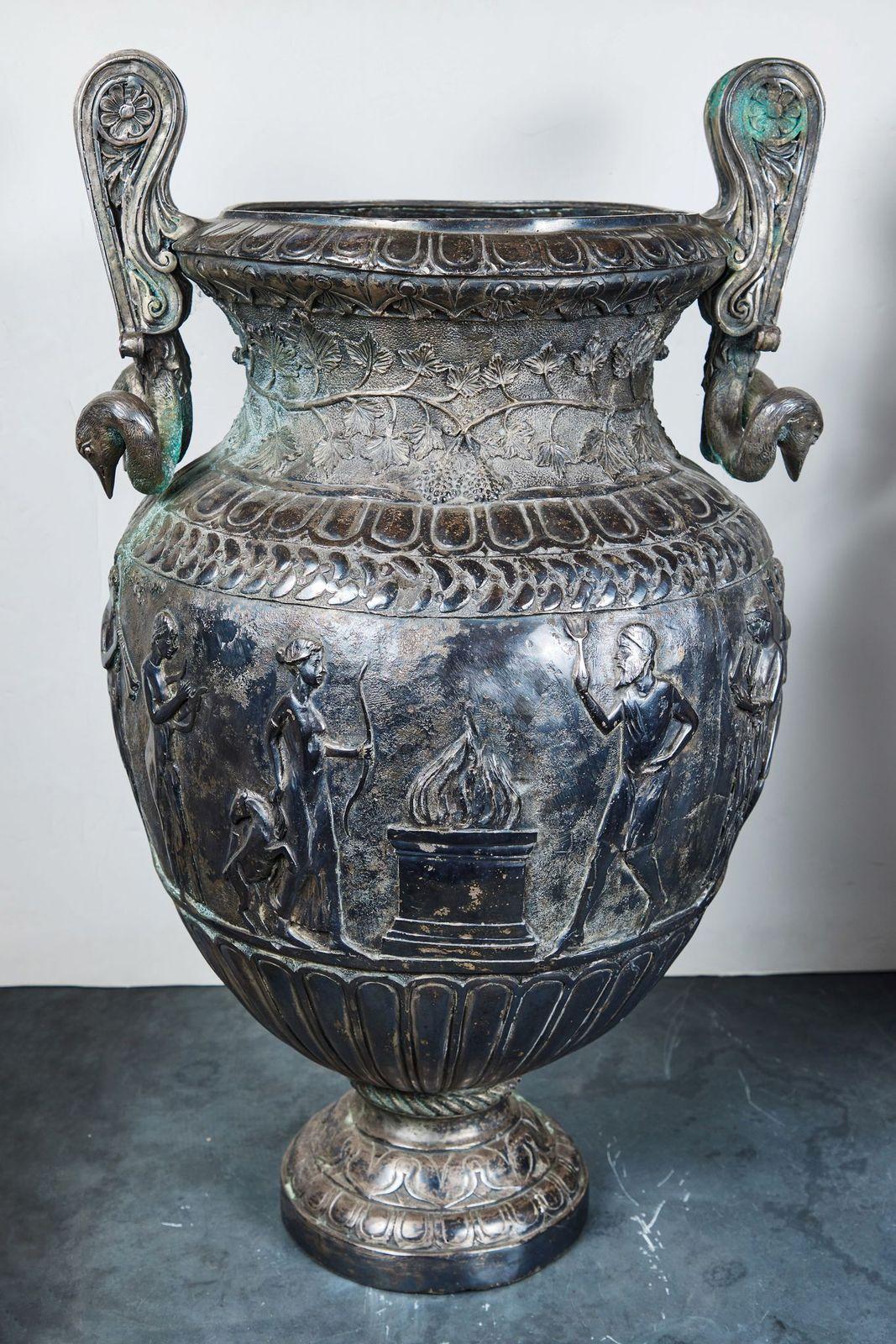 A glorious pair of c. 1915, Italian, silvered metal Neoclassical revival urns featuring a central band of celebratory relief figures proceeding toward a ceremonial fire. The top boasts impressive, dual swan-head handles that cascade toward main