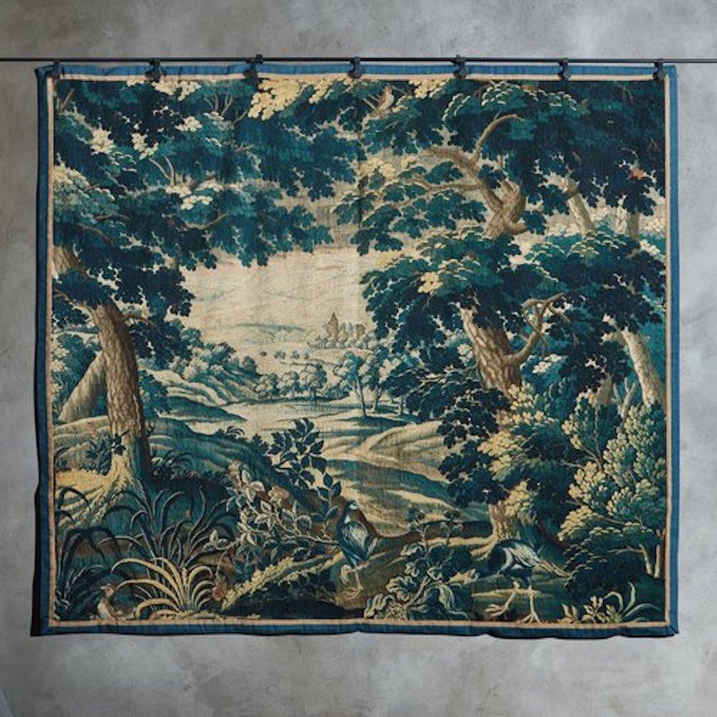 A large scale early 20th Century Belgian tapestry featuring a lush verdant woodscape design with bird motifs in rich blue, green and taupe hues. It has a navy border, a cream backing and beautiful time honored wear. Sourced in Belgium, 20th Century.