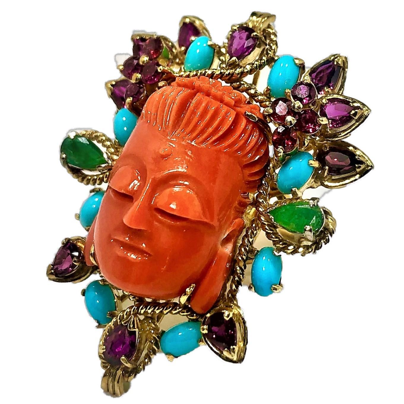 An exotic 14K yellow gold ring, featuring a finely carved coral Buddha's head measuring approximately 24mm X 16mm. The face is surrounded by oval cabochon cut turquoise, pear shaped emeralds, along with pear shaped and round cut purple/pink