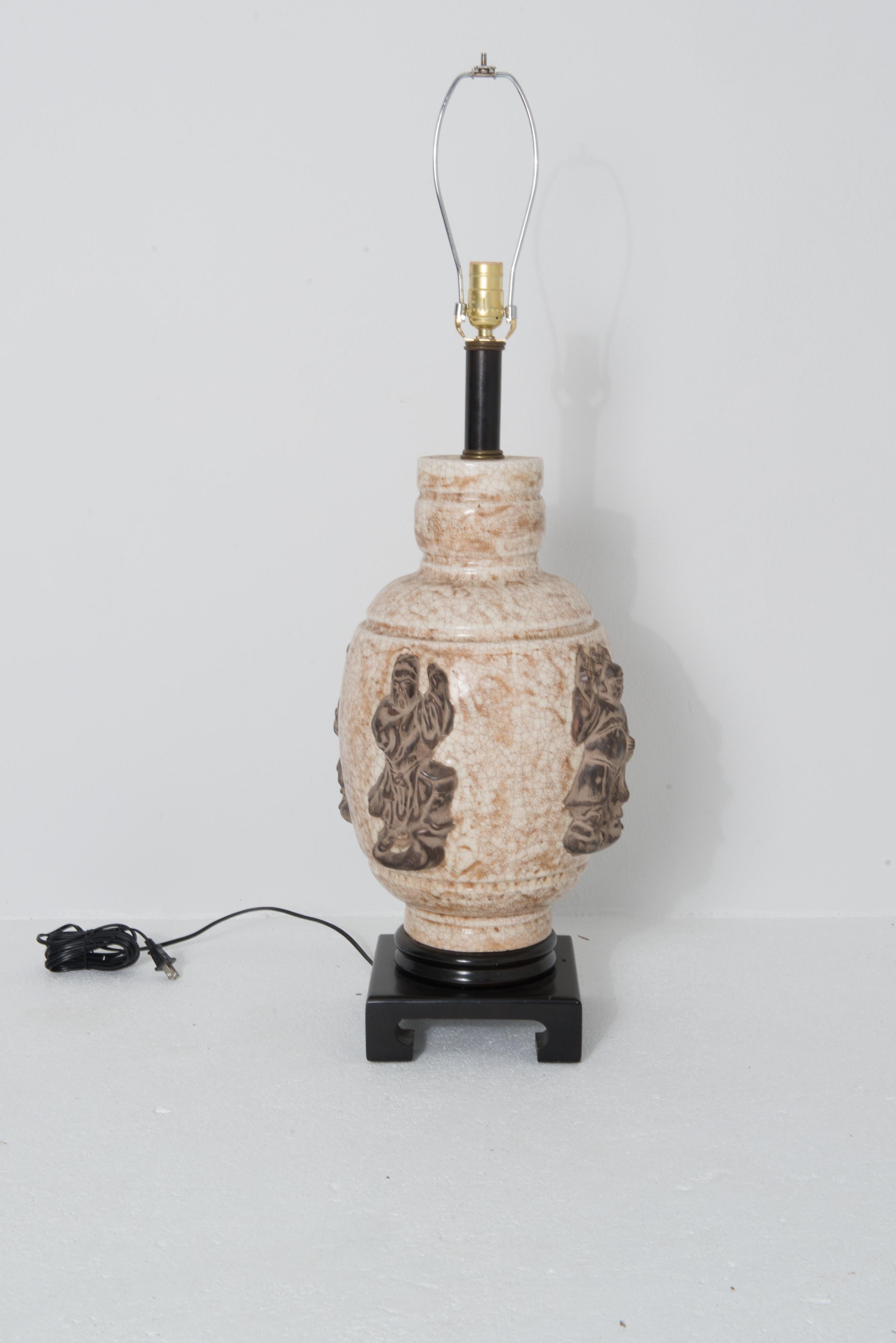 Eine großformatige Keramiklampe im asiatischen Stil in Creme- und Sandtönen, die mit vier großen, hochreliefierten, zweifarbigen braunen Figuren verziert ist. Die geschnitzte Holzbasis ist 8