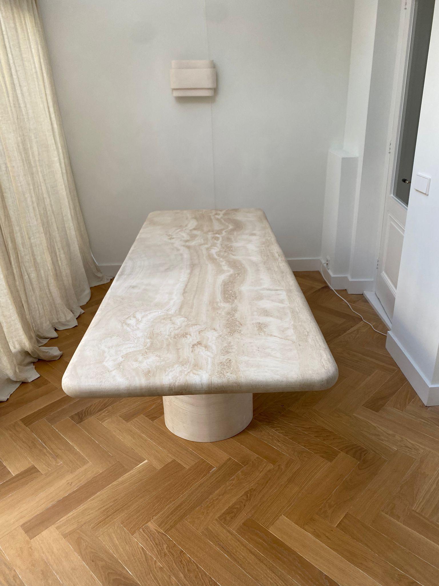 Eine funktionelle Skulptur des Raumes. Dieser organisch-moderne, minimalistische Tisch aus getrommeltem Travertin hat eine dicke Tischplatte mit geschwungenen Kanten, die elegant auf einer geschwungenen zylindrischen Säule ruht. 

Die markante
