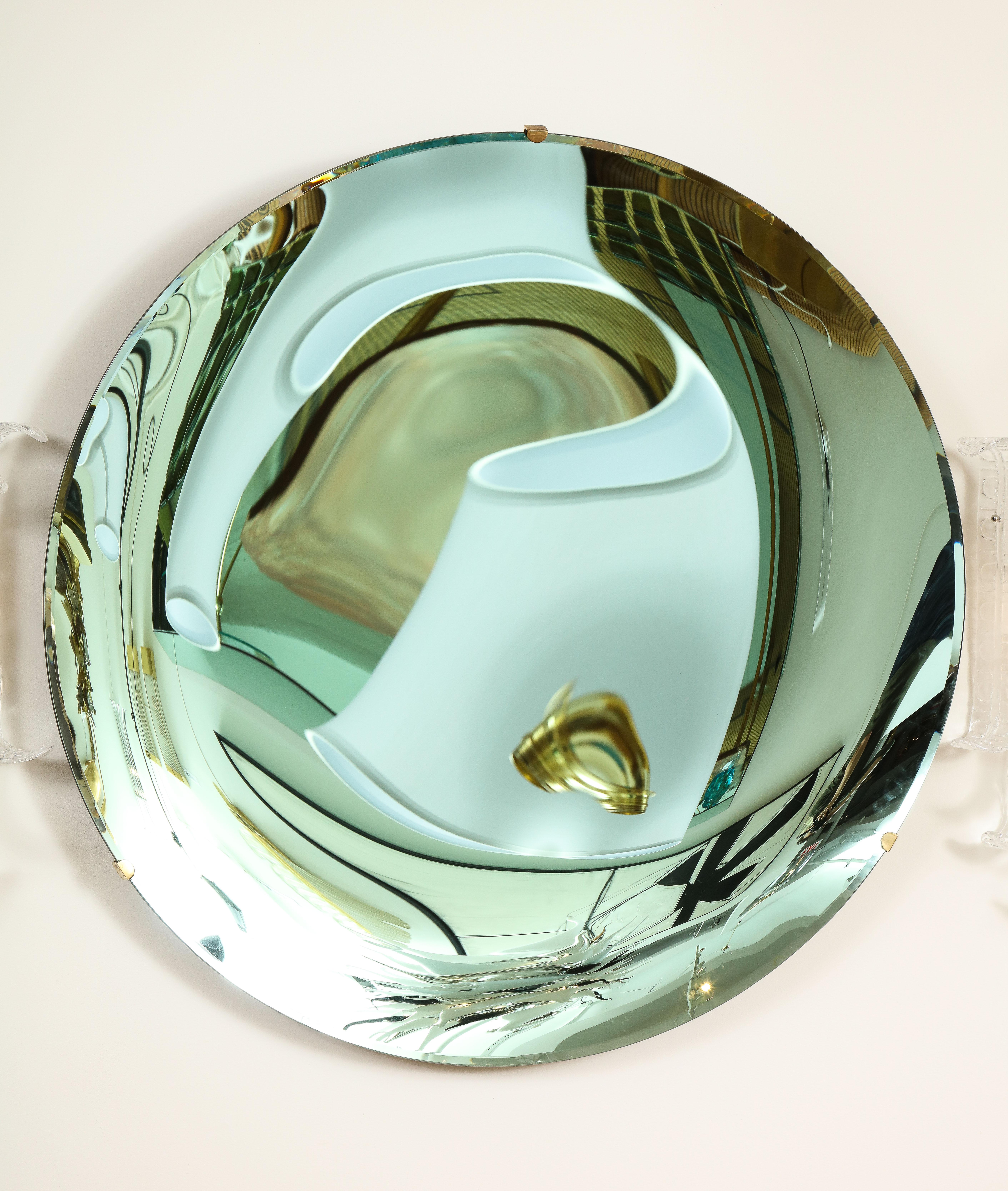 Large round tinted glass in a beautiful soft green hue, thermoformed into a sculptural concave form and mirrored. Mounted on a brass structure that is attached to wall. This mirror is a sculptural art piece that changes hues depending on its angle