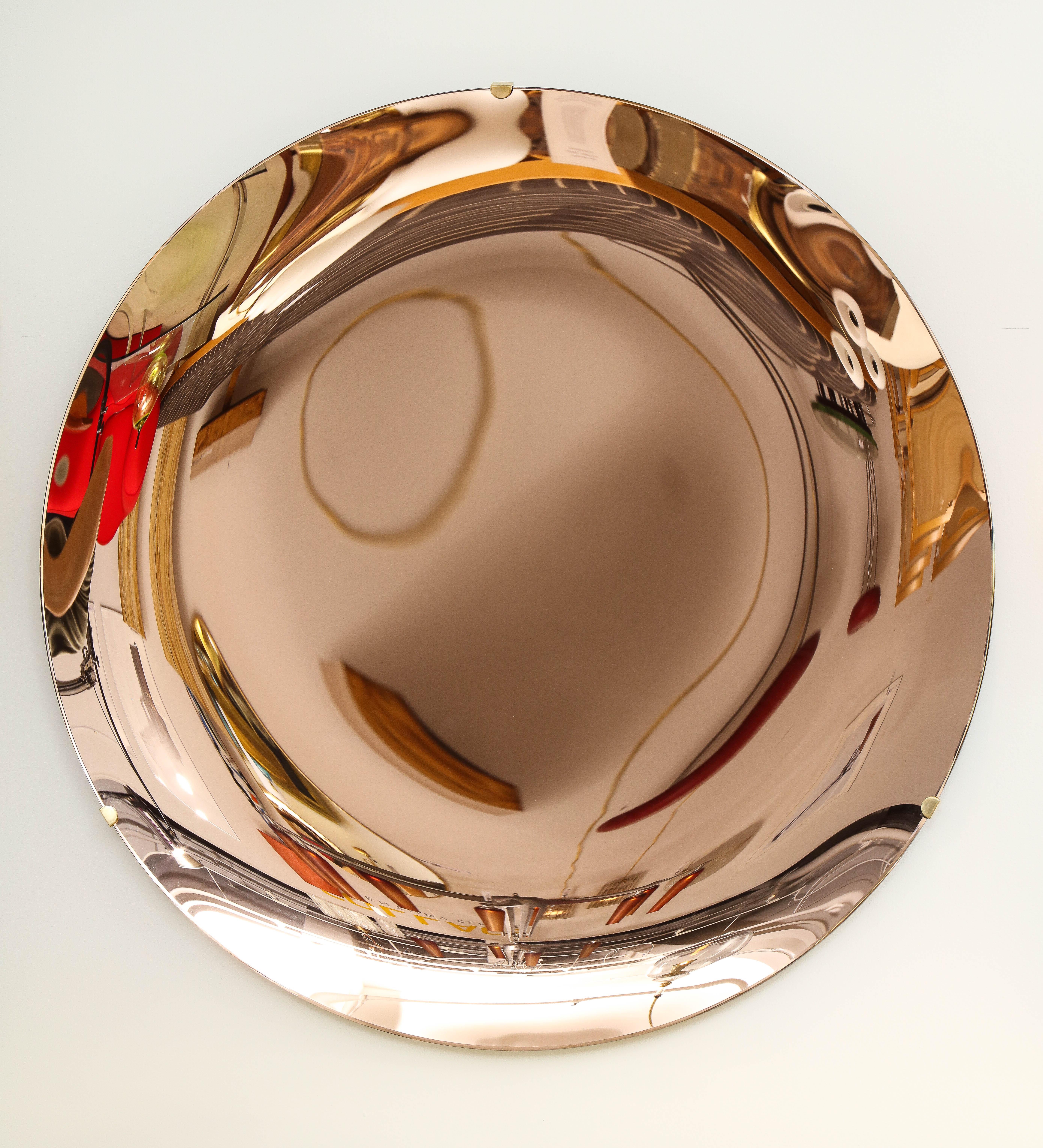 Large round tinted glass in a beautiful soft salmon or rose hue, thermoformed into a sculptural concave form and mirrored to create a stunning optical lens effect. Mounted on a brass structure that is attached to wall. Hand-casted in Milan, Italy,