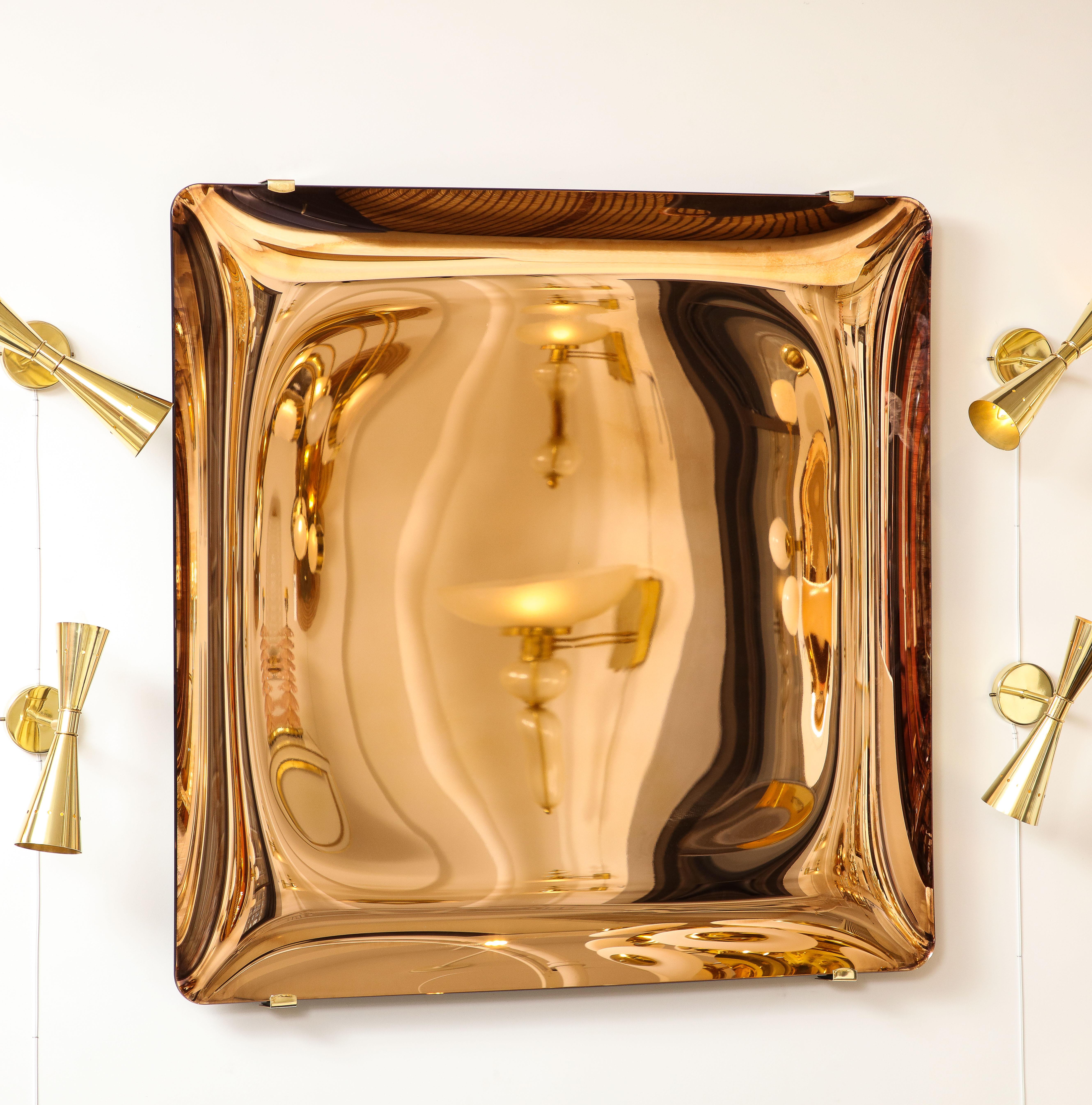Large square frameless tinted glass in a rose gold color, thermoformed into a sculptural convex form and mirrored on back. Mounted on a steel with brass four arm structure that is attached to wall. Only brass tips show on front. Hand-casted in